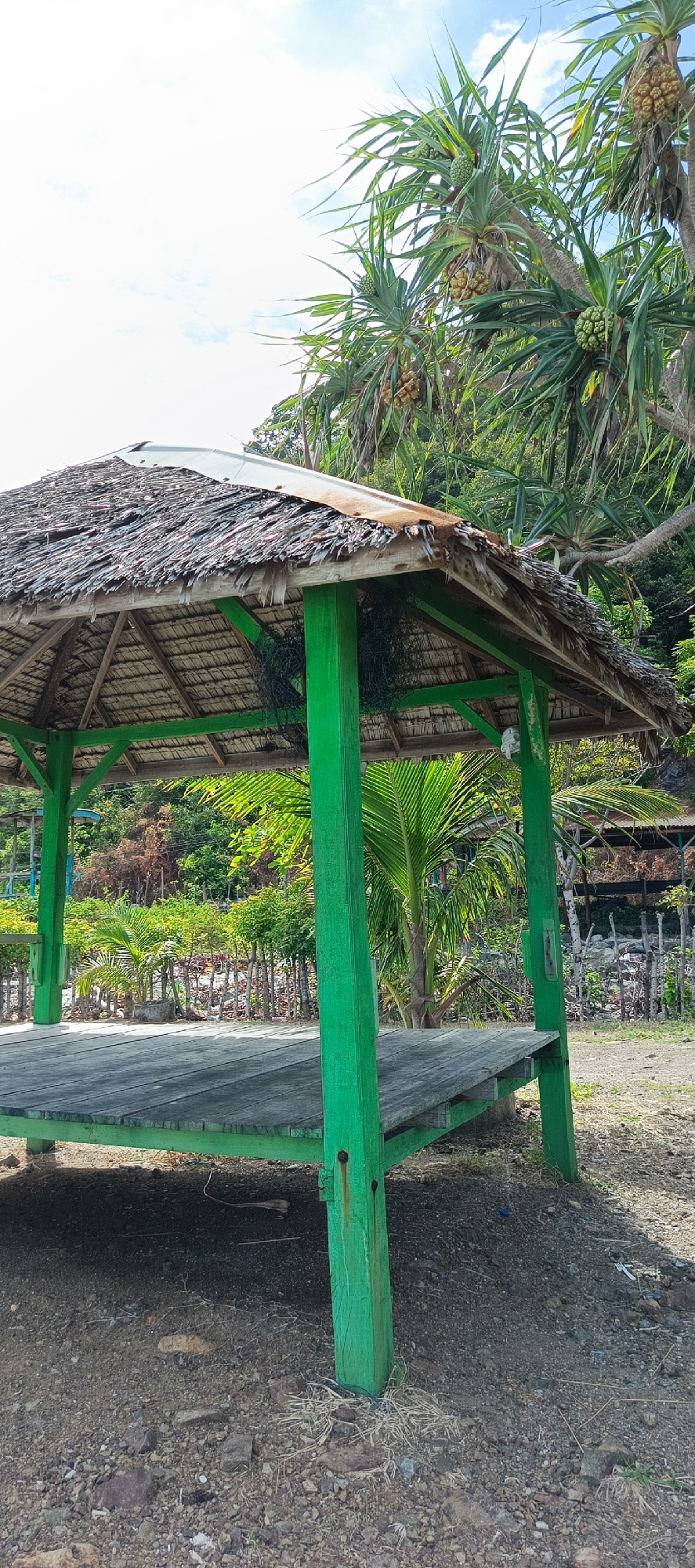 An open-air, wooden gazebo with a thatched roof stands on a gravelly ground surface. The gazebo's four corner posts are painted bright green, and the thatched roof appears weathered, with some sections of corrugated metal visible underneath. A wooden platform forms the floor of the gazebo. In the background, lush green vegetation, including palm trees and other tropical plants, covers a hillside. The sky above is partly cloudy. The scene is located in Naga Umbang, Indonesia, suggesting a tropical coastal or hilly environment. No people are present in the image.