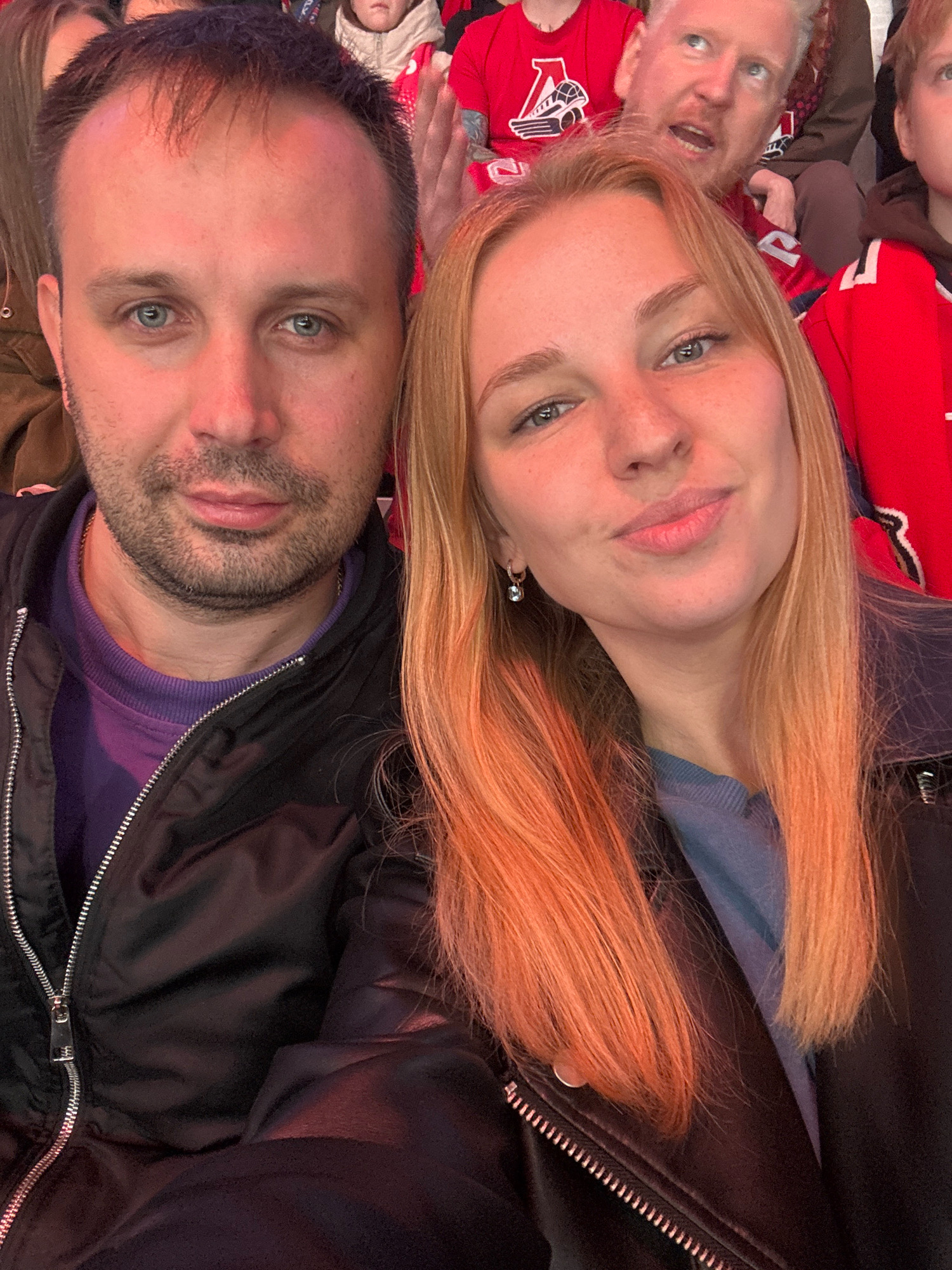 In this medium shot, a Caucasian couple is positioned in the foreground, their faces slightly turned towards the camera in a selfie. The man, on the left, has short, dark hair, light blue eyes, and stubble. He is wearing a purple sweatshirt under a black zipped jacket. The woman, on the right, has long, blonde hair with orange undertones and blue eyes. She is wearing a light blue top and a black jacket, with a small hoop earring in her visible ear. Behind them, other individuals are visible in a seating area, likely a stadium or arena, indicated by red sports jerseys worn by some spectators. The overall lighting is warm, suggesting an indoor setting. The image does not contain any recognizable landmarks or clues to definitively place the location in Rusanovo, Russia.