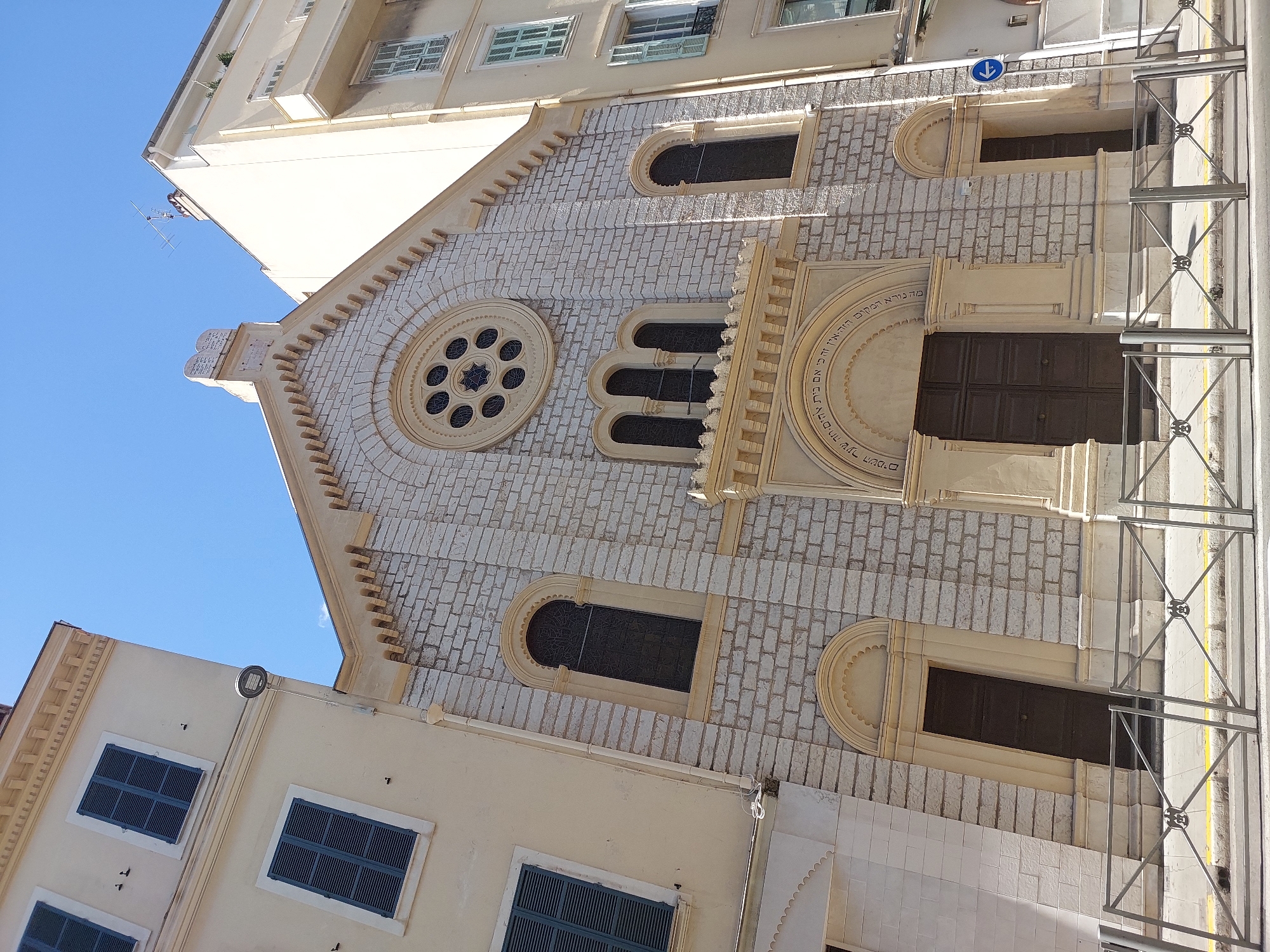 The image shows a street view of a synagogue in Nice, France, under a clear blue sky. The synagogue features a light-colored stone facade with a large rose window at the top, adorned with a star of David. Below the rose window are three arched windows, and a decorative inscription in Hebrew is visible above the main entrance. The building is flanked by a pale yellow building with dark blue shutters on the left and another taller, pale yellow building with green shutters on the right. A metal fence with decorative elements separates the sidewalk from the street, where a blue sign with a white arrow points left. The scene is bathed in bright sunlight, casting shadows on the buildings and the street.