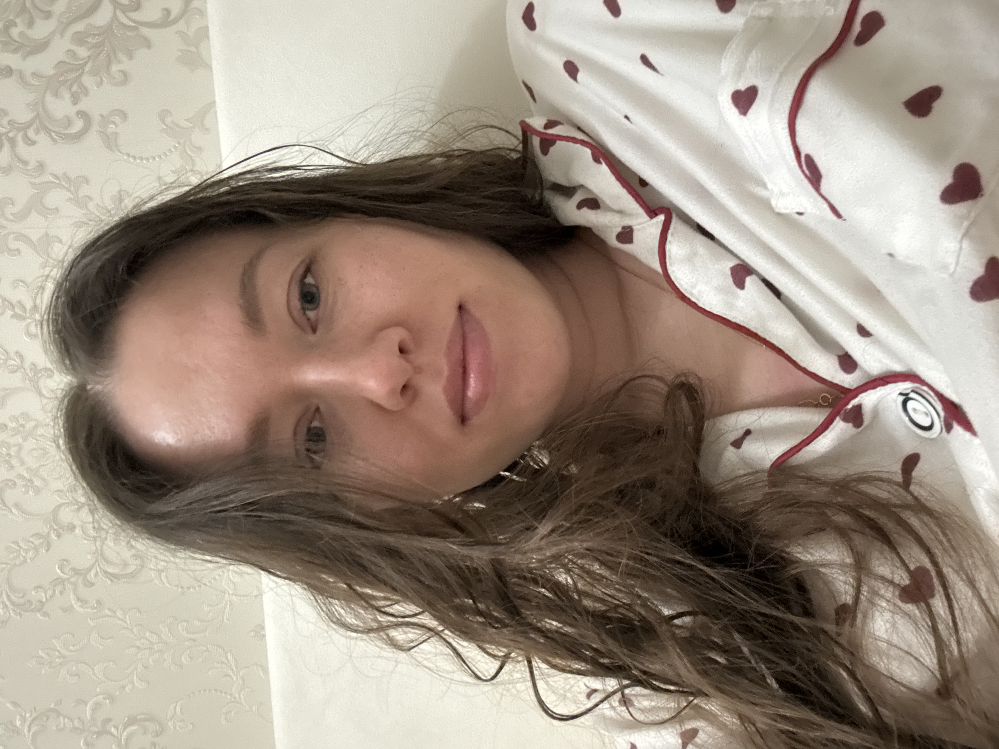 A woman with long, wavy brown hair lies on a bed covered in white floral patterned linen. She is wearing a white pajama top with red heart designs and red piping. She is fair-skinned with light eyes and light pink lips. The photo appears to have been taken indoors in a personal space. There is no evidence to suggest this is Ferma, Russia.