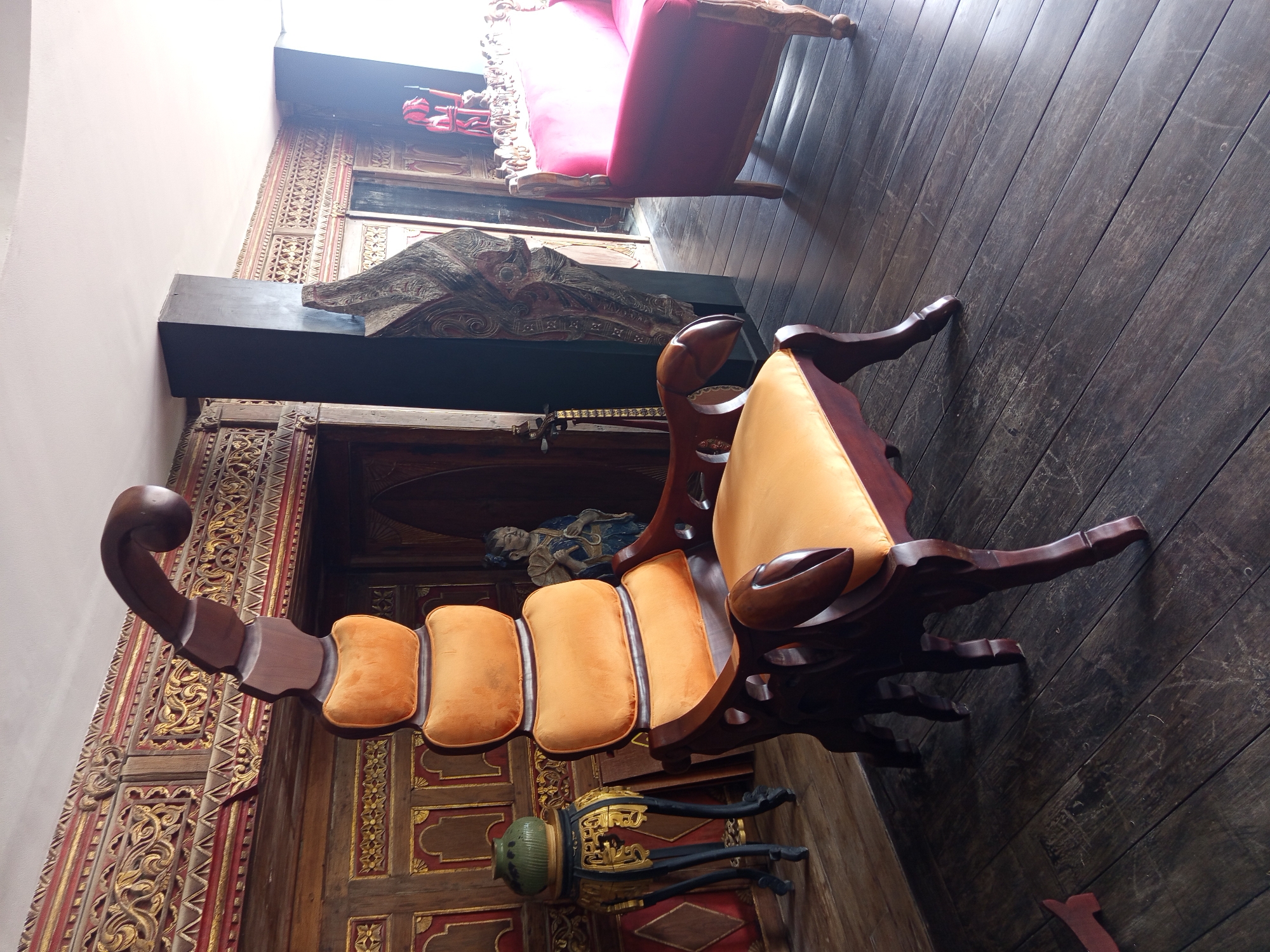 A brightly lit, indoor photograph taken in North Kuta, Indonesia shows two ornate, antique chairs. The chair in the foreground is particularly striking. It is shaped like a scorpion, with the tail serving as the backrest. The cushions are bright orange, contrasting with the dark wood frame. The armrests are carved into claw shapes. Another chair, upholstered in red velvet, is partially visible in the background. The floor is made of dark, polished wood planks. An elaborately carved wooden wall with gilded details is visible to the left of the scorpion chair, while a dark, wooden shelf with carved figures sits above a doorway in the background. A small, ornate table with a green vase stands below the scorpion chair.
