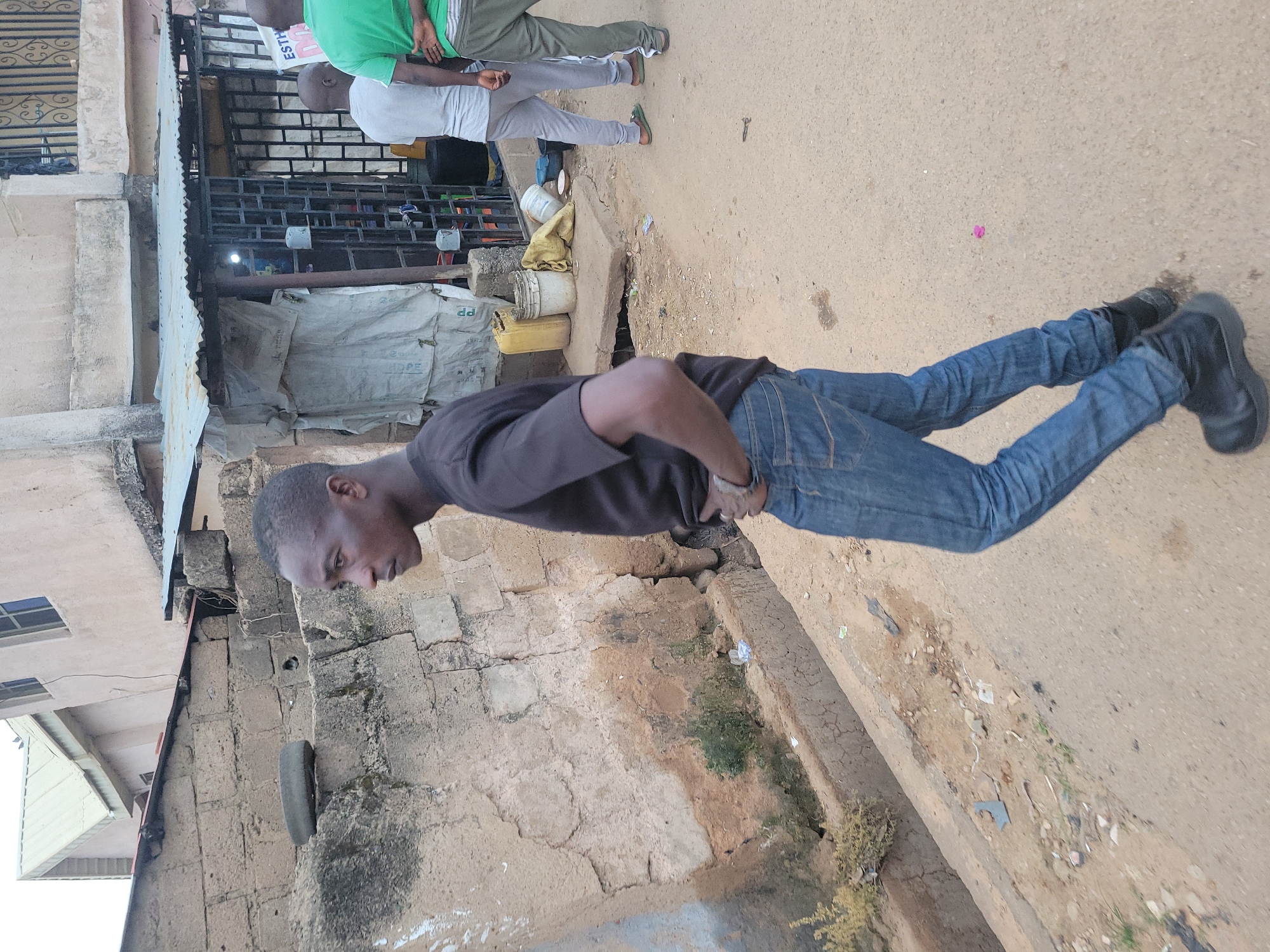 A dark-skinned man in a black t-shirt and blue jeans walks on a dusty road in Jos, Nigeria. He is seen from the side, with his left hand in his pocket and his right hand by his side. Behind him, a small storefront with a corrugated metal roof is visible, with several people standing in front of it. The storefront appears to be selling various items, including buckets and containers. The area is somewhat dilapidated, with crumbling walls and debris on the ground, indicative of a developing urban environment. A drainage ditch runs along the side of the road, partially filled with water and debris. The overall impression is that of a busy street scene in a West African city.