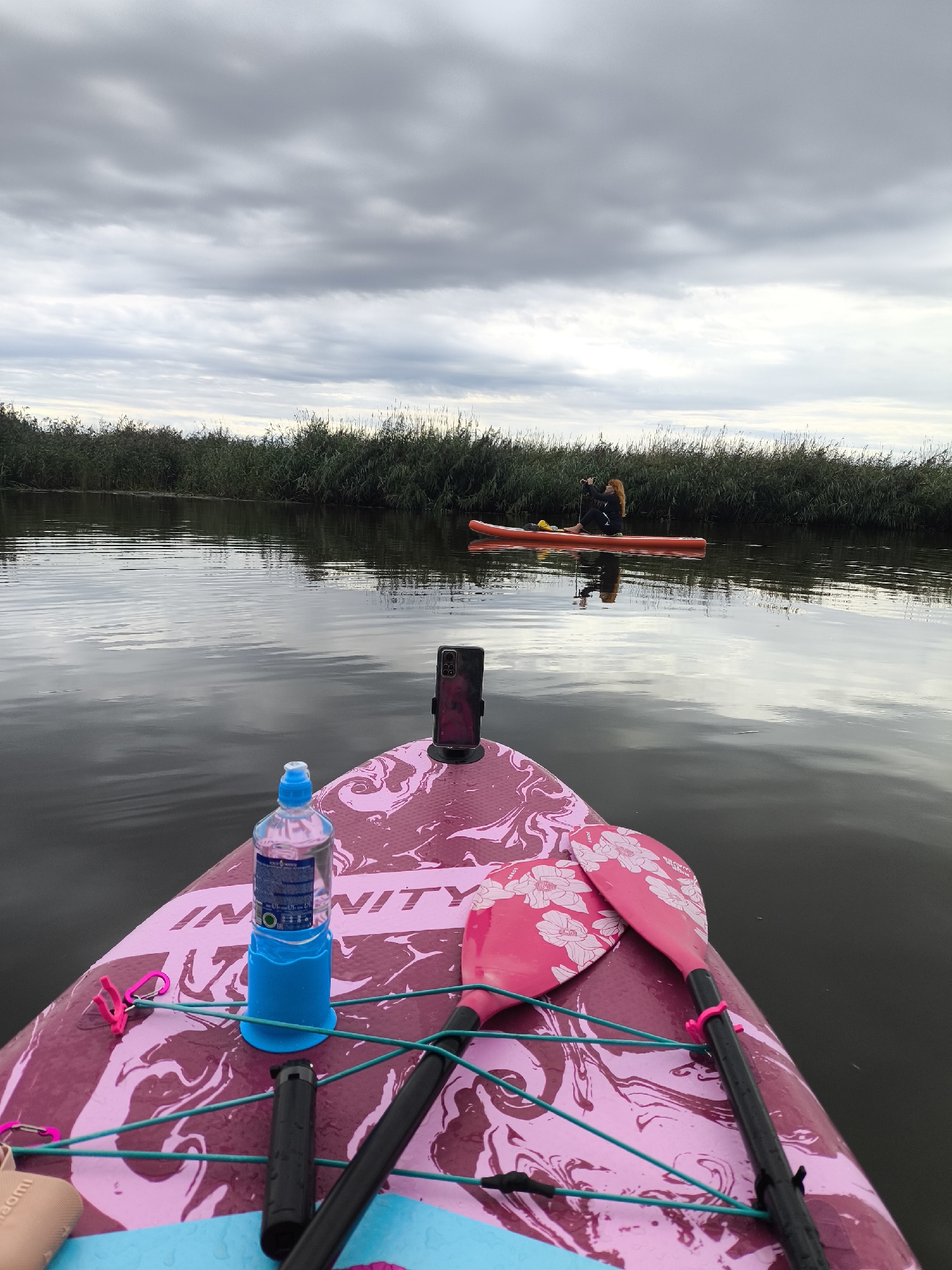 The image shows a perspective from a paddleboard on a calm body of water, likely a river or canal, with dense reeds lining the far bank. The sky is overcast with gray clouds. In the middle distance, another paddleboarder with red hair is seated on an orange paddleboard, facing away from the camera and paddling. The foreground of the image is dominated by the pink and marbled patterned surface of the paddleboard from which the photo is taken. Visible objects on this paddleboard include a blue bottle holder with a water bottle, a black smartphone mounted on a stand, and two pink-blossomed paddleboard paddles. Two pink carabiners are attached to turquoise bungee cords. The location is identified as Ushakovo, Russia.