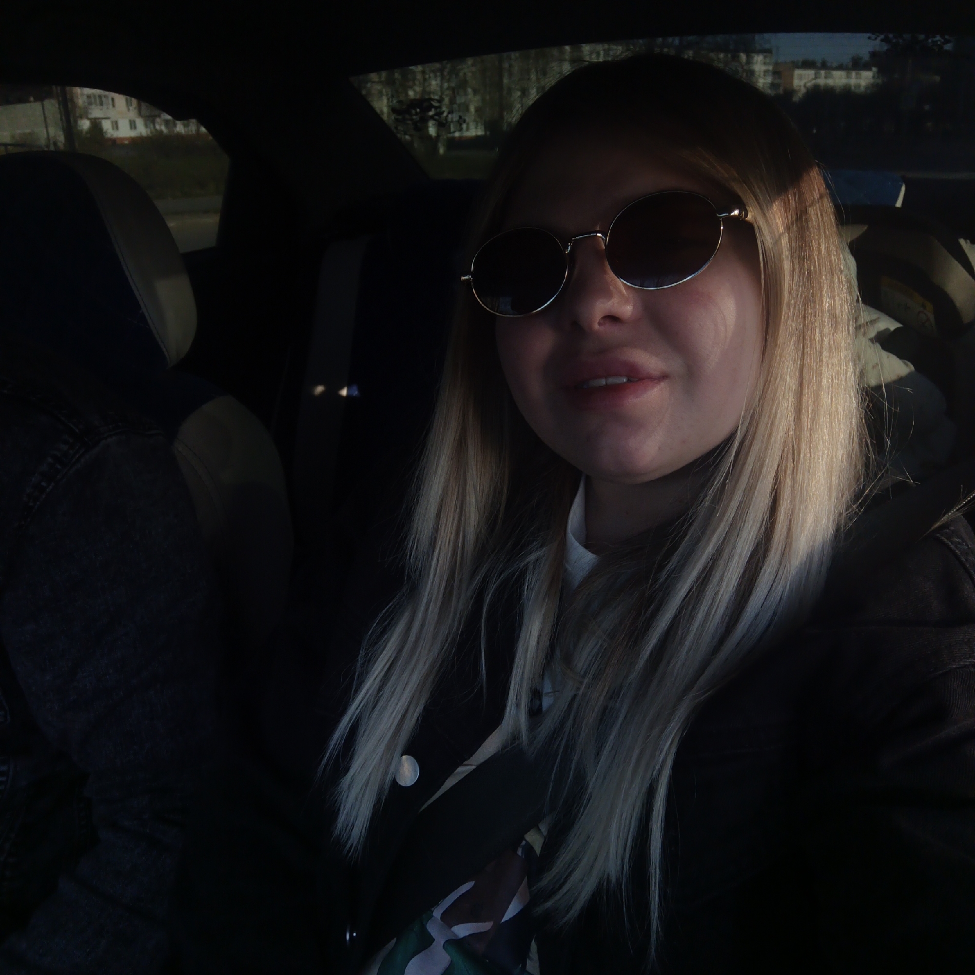 The image shows a woman wearing sunglasses and a jacket, sitting in the passenger seat of a car. The background is blurred, but appears to show buildings, indicating an urban environment. The specific city location is unknown.