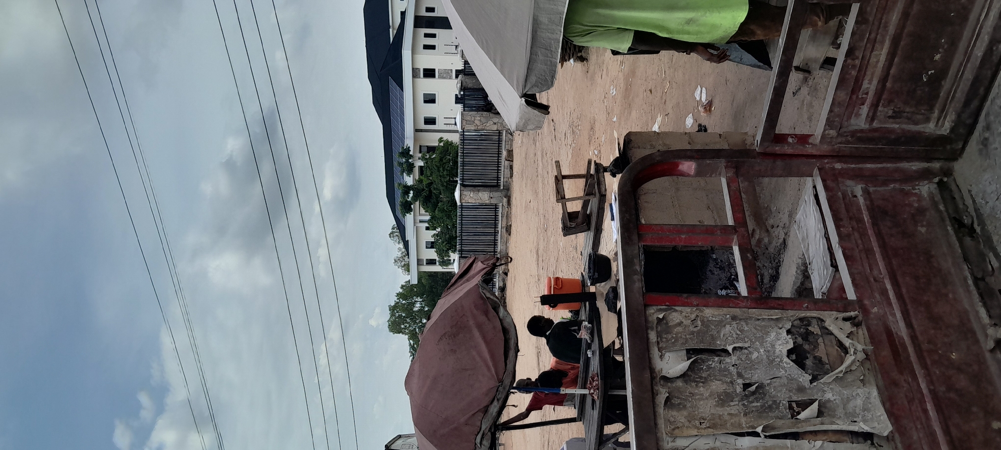 The image depicts an outdoor scene in Jalingo, Nigeria. In the foreground, a weathered, red metal structure with peeling paint is prominent, suggesting a food stall or preparation area, with visible charcoal embers inside. Behind this, a wooden table is covered by a brown tarp, providing shade for two individuals. One person, wearing a red shirt, is partially visible, and the other, in a dark shirt, is seated. Food items, a black pot, and an orange cooler are on the table. To the right, a light-colored umbrella casts shade over a person in a green shirt, partially obscured. Further back, a modern building with solar panels on its dark roof is visible behind a black metal fence and stone pillars. Trees line the background, and the ground is sandy. The sky is overcast with scattered clouds.