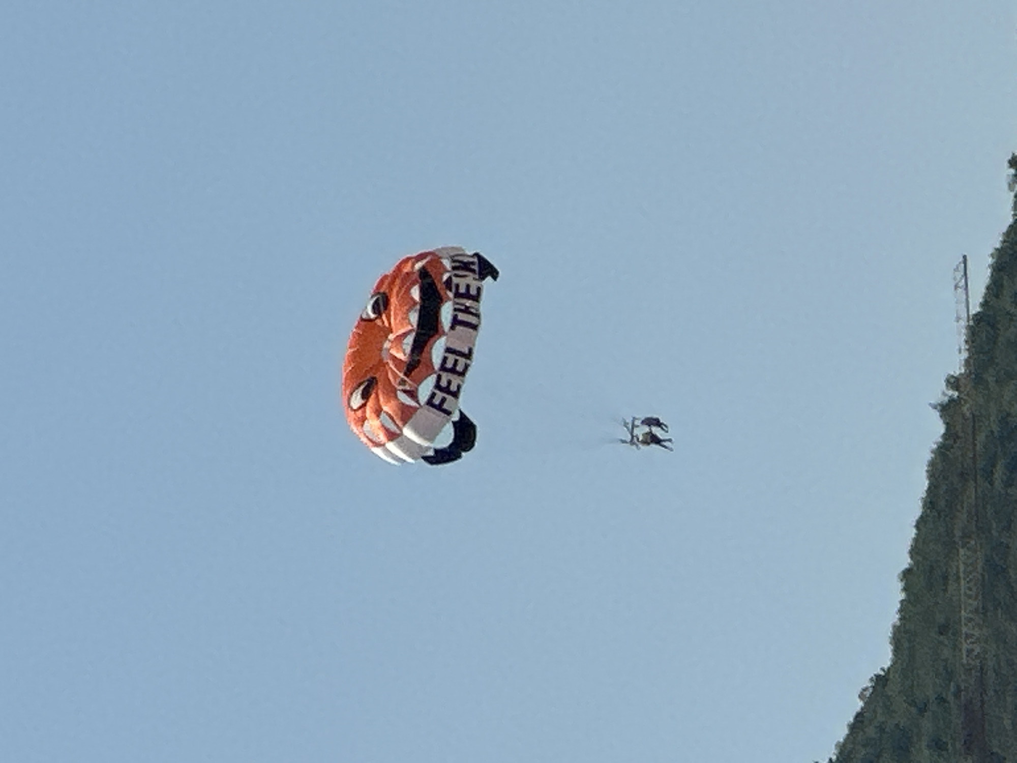 An orange and white parasail, designed to resemble a jack-o'-lantern face with black eyes, a grinning mouth, and white teeth, is visible in a clear blue sky. The words "FEEL THE W..." are printed in black on the lower portion of the parasail. Attached to the parasail by ropes, two individuals are parasailing. Below the parasail and individuals, a green, tree-covered hillside is visible. This scene is indicative of a tourist activity, likely parasailing, taking place over the water near Budva, Montenegro.