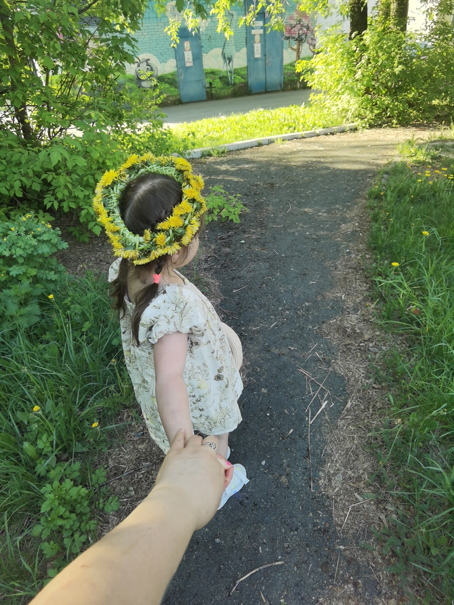 A person holds a young child's hand, leading the child down a narrow asphalt path. The child wears a dress with a floral pattern and a wreath of yellow dandelions on her head. The path is bordered by green grass and foliage on both sides. In the background, a blue building with graffiti can be seen. This image was taken in the city of Perm, Russia.