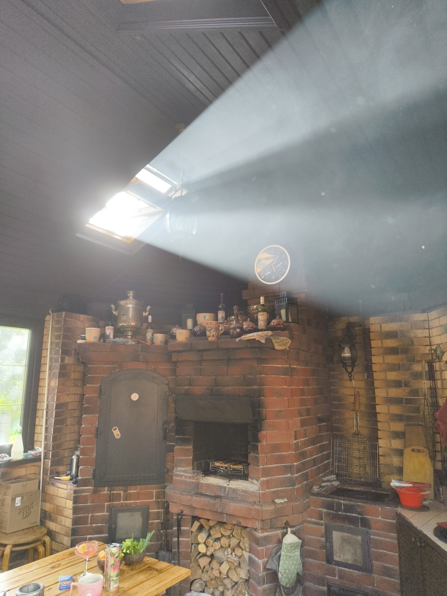 The image depicts an outdoor covered patio or veranda area, likely in a rural setting, possibly a dacha in Востец Снт, Russia, given the context provided. The main subject is a large, ornate brick barbecue and oven structure that dominates the right side of the frame. This structure features a main oven cavity with a metal door, a smaller compartment below it, and a grill area to the right, where cooking implements like a grill basket are visible. The brickwork shows signs of use, with soot and charring around the cooking and oven areas.
Above the oven structure, a mantelpiece or shelf displays a collection of items, including a traditional Russian samovar, several cups or mugs, bottles, and decorative pieces. A clock with a circular face is mounted on the brick wall above the shelf, to the right of the samovar.
The ceiling of the patio is dark-colored, possibly wood or metal, with visible beams or slats. A skylight is positioned in the ceiling, allowing natural light to enter. A strong beam of sunlight cuts diagonally across the upper portion of the image, illuminating dust particles in the air and creating a dramatic effect.
In the foreground, a wooden table is set with a few items: a pink mug, a beverage can, a glass with a drink, a small bowl of green vegetables, and some packets on a blue package. To the left of the table, a wooden stool is visible, with a cardboard box on top.
The environment appears to be a backyard or garden area, with greenery visible through a window on the far left. The overall impression is one of a rustic, functional outdoor living space, possibly used for gatherings and cooking. No people are visible in the image.