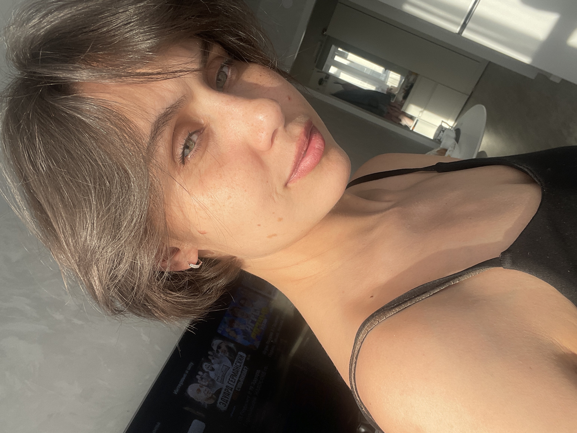 A close-up, slightly angled photograph captures a light-skinned woman with short brown hair. She is wearing a black tank top. She has light eyes and her lips are lightly colored. The background is a modern interior, including a TV screen and a glimpse of a kitchen area with white cabinets. The image is taken in a sunny setting, evident from the strong light source. The specific location in Izhevsk, Russia, is not identifiable from this image.