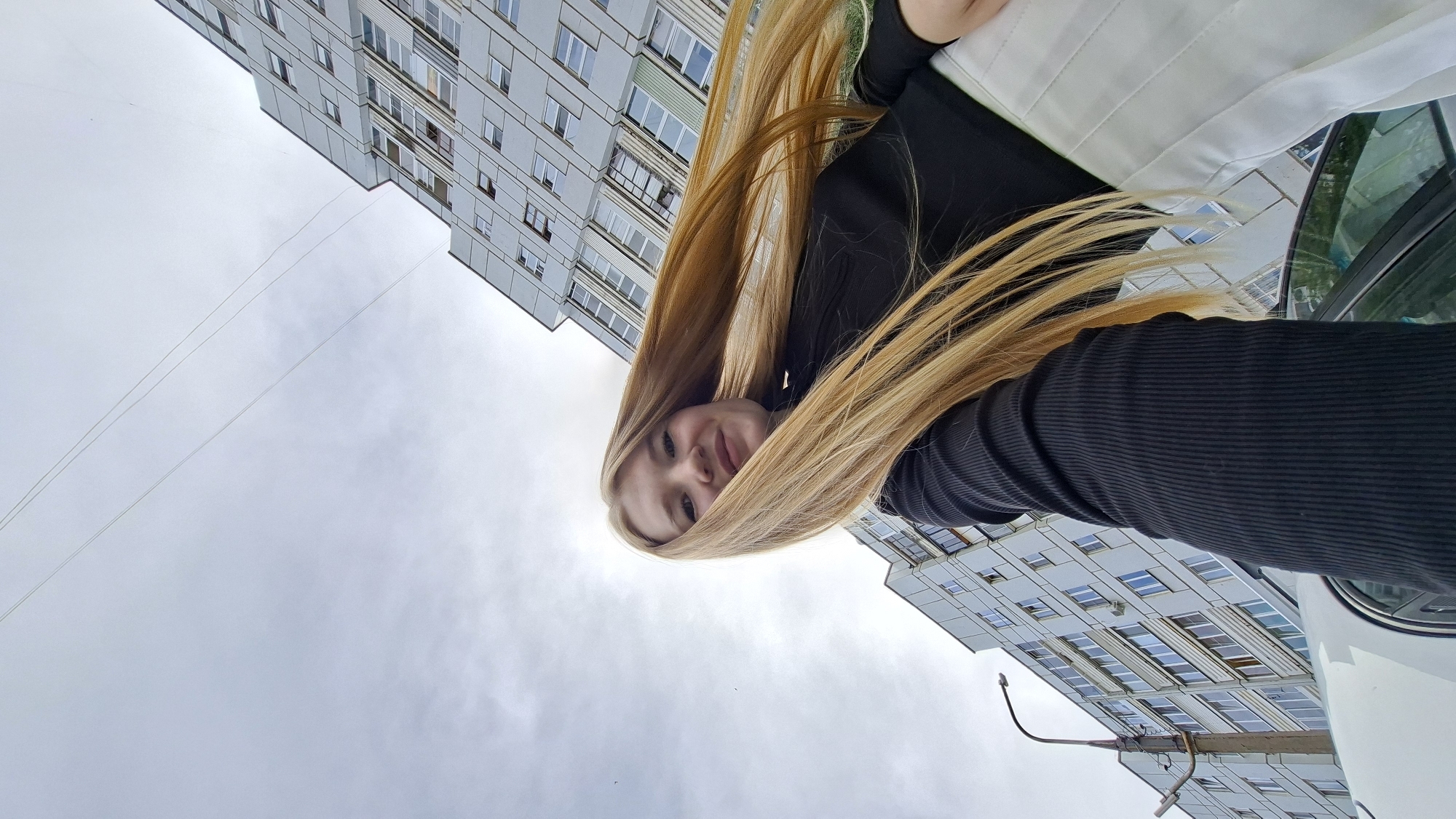 A young woman with long, blonde hair is captured in a low-angle, outdoor photograph. She is wearing a black, ribbed, long-sleeved top and a white skirt. Her hair is blowing dramatically to the right, framing her face and body. She is smiling directly at the camera. In the background, a modern, multi-story building with many windows dominates the upper right portion of the frame, with another building visible on the left. A street lamp is also visible on the left side of the image. The sky is overcast, with visible power lines stretching across it. The photograph appears to have been taken in the city of Chaikovsky, Russia, given the architectural style of the buildings and the overall urban setting. A white car is partially visible in the foreground.