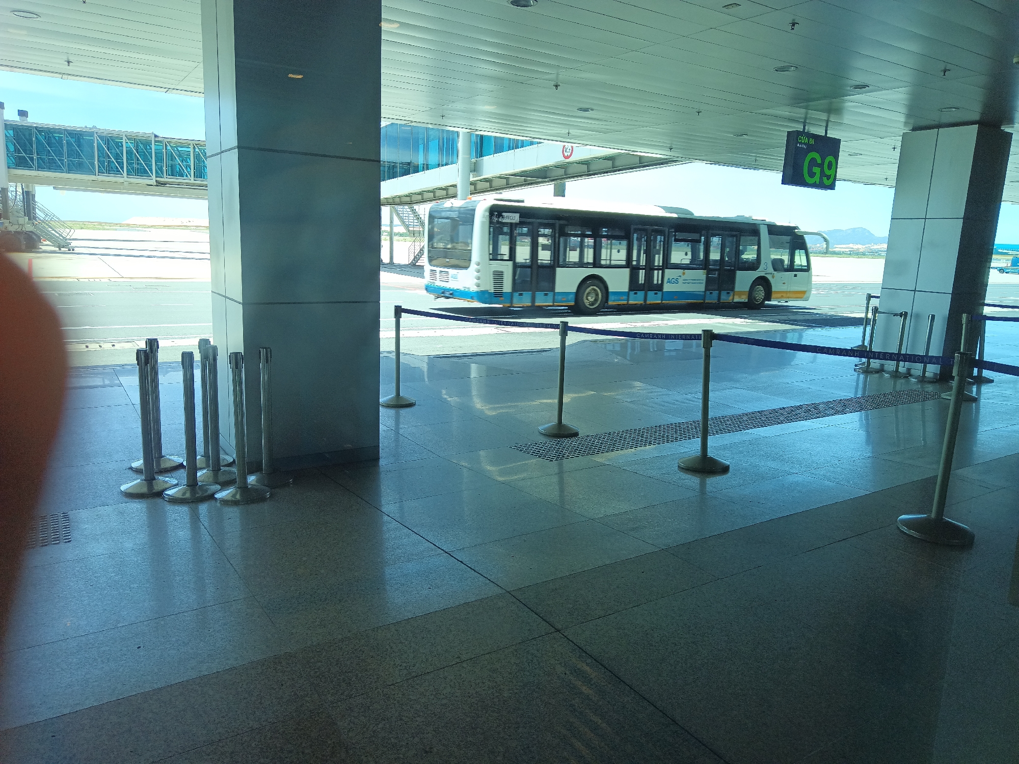 This image captures a scene at Cam Ranh International Airport in Bắc Cam Ranh, Vietnam. A white and blue airport bus, labeled "AGS" and with "NHATRANG" visible above the windshield, is moving along the tarmac. In the background, a jet bridge extends from a terminal building, and mountains are visible on the horizon. The foreground shows a modern airport terminal walkway with polished floors, metal stanchions with ropes, and a sign indicating "G9". The lighting suggests it is daytime with bright sunlight casting reflections on the floor.