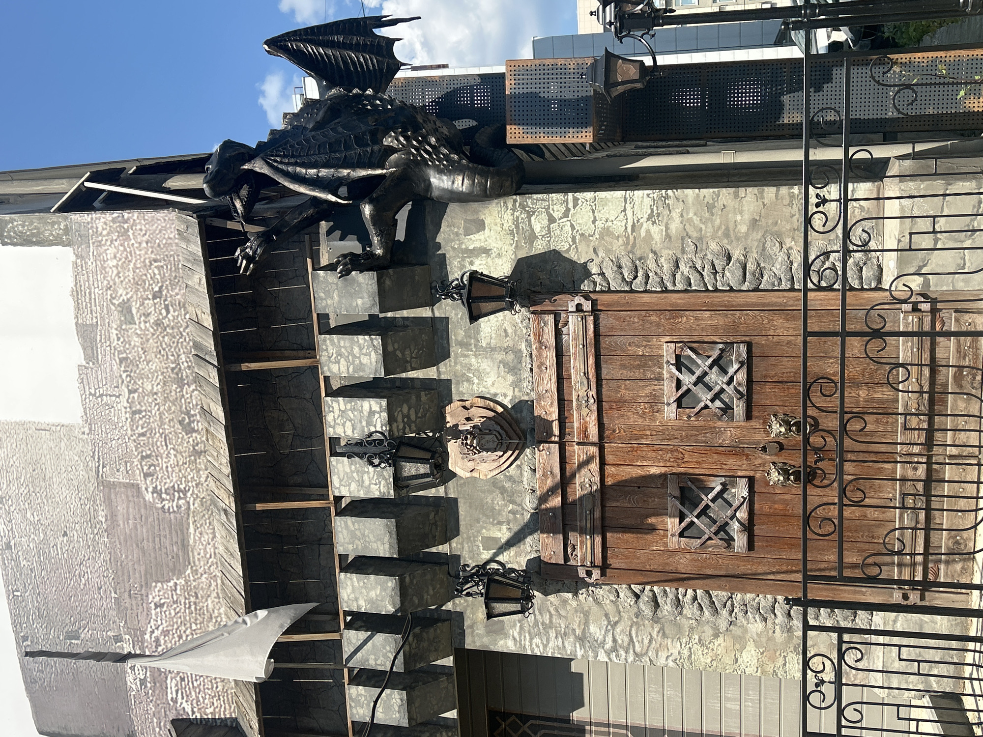 This is a medium shot of an ornate, rustic building facade, possibly a facade for a shop or restaurant, with a clear blue sky and scattered clouds above. A large, dark, metal dragon sculpture is perched on the roofline, its wings spread and mouth open. The building's exterior is a mix of textures and materials. A large, rustic wooden door with two small, square windows with decorative metal grates is prominent. Above the door, the wall is textured to resemble rough stone. Several wrought-iron lanterns hang from the building, casting shadows. A decorative black wrought-iron fence with swirling motifs encloses part of the area. To the left, a large, white tarp or sail-like structure is draped over a wooden frame, with a textured mural visible behind it. The overall impression is one of fantasy and medieval-inspired design. The lighting suggests it is daytime, likely late morning or early afternoon, with bright sunlight. The location is identified as Perm, Russia. There is no visible text in the image.