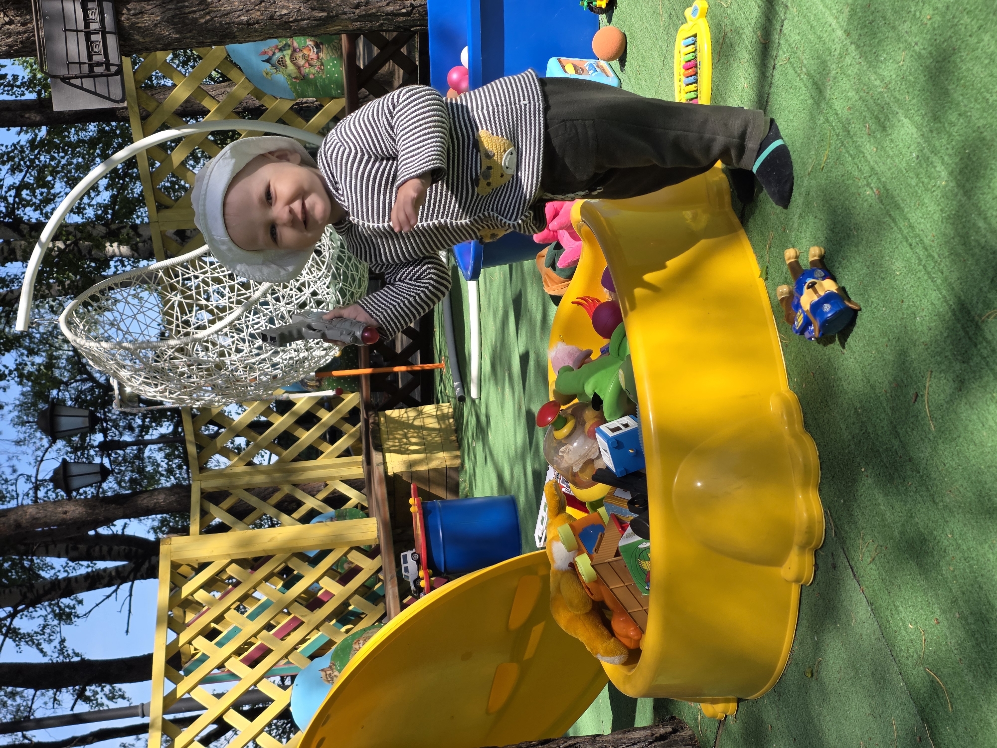 Here's a detailed description of the image:
**Location:** The photograph appears to be taken in an outdoor play area, possibly in the city of Chaikovsky, Russia, based on the user's prompt.
**Subjects:** The primary subject is a young child, likely an infant or toddler, wearing a white hat and a striped shirt. The child is standing near a large yellow toy bin.
**Objects:** Key objects in the scene include:
* A large, yellow toy bin filled with various toys.
* Toys strewn on the green artificial turf, including a blue Paw Patrol figure.
* A hanging white chair.
* A yellow latticework structure.
* Other playground equipment and toys visible in the background.
**Actions/Interactions:** The child is holding a toy gun and appears to be playing near the toy bin. The exact nature of their play is not possible to determine with certainty.
**Environment:**
* The scene is set outdoors on a bright day with natural sunlight.
* The ground is covered with green artificial turf.
* There are trees visible in the background, suggesting a park or yard setting.
* The environment seems to be well-maintained and designed for children's play.