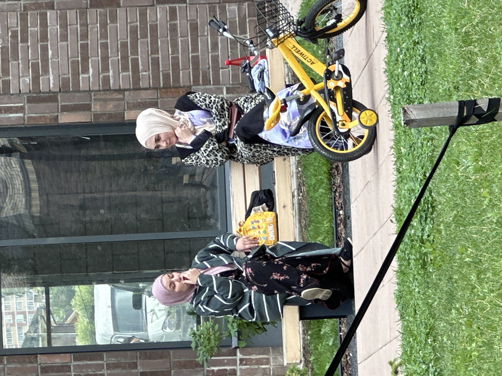 Two women, both wearing hijabs, are seated on a wooden bench. The woman on the left wears a green and white striped cardigan over a dark floral dress and a pink hijab. She is leaning forward with her hand near her mouth, appearing to be speaking or thinking. On the bench next to her is a yellow bag with cartoon characters and the text "AMONG US" visible. The woman on the right wears a leopard print abaya over a light-colored hijab and a white and purple patterned garment. She is resting her chin on her hand, looking to her left. In front of the bench, a bright yellow children's bicycle with training wheels is parked. The bicycle has a basket on the front and the word "ACTIWELL" printed on its frame. Behind the bench is a building with large windows reflecting an outdoor scene, including a white van and trees. The ground in front of the bench is paved with light-colored square tiles, and further in the foreground, green grass is visible with a wooden post and a black cable. The scene appears to be in an urban outdoor setting, potentially a residential complex or park area, consistent with the described location in Znamya Oktyabrya, Russia.