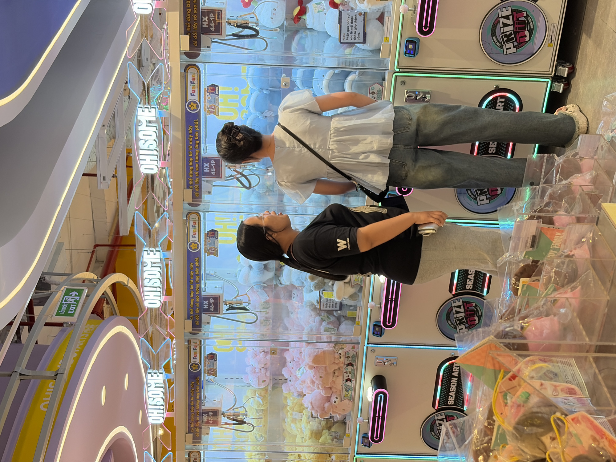 Two young women stand in front of claw machine games at a shopping mall. The image is likely taken in the city of Thủ Đức, Vietnam. The woman on the left wears a black T-shirt with a white "W" and gray shorts. The woman on the right wears a light blue shirt, dark jeans, and a black bag across her shoulder. The claw machines have plush toys inside and signs in Vietnamese, including the words "OH!SOME," "FuniFun!," "EXIT," and "PRIZE OUT." There are also acrylic display boxes full of toys nearby.