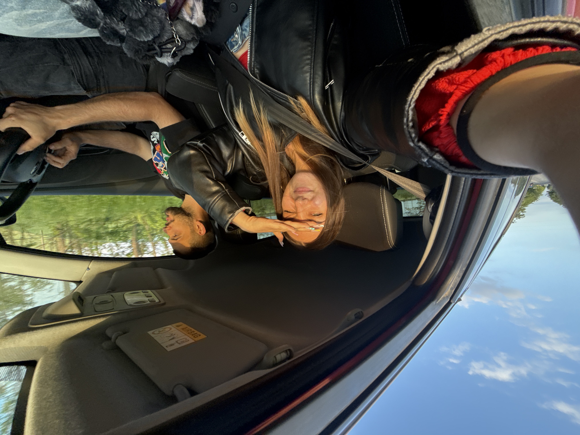 A selfie taken from inside a vehicle shows a woman wearing a black leather jacket, secured with a seatbelt. Her right hand is raised to shield her eyes. A man is visible in the driver's seat. The car's interior includes a sun visor with a warning label. The scene suggests daylight, with a partial view of trees through the window. The location in Babovo, Bulgaria is not evident in this image.