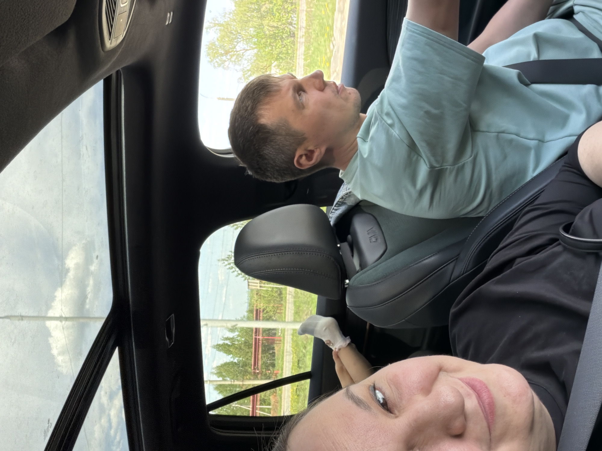A photograph taken inside a car, with a woman in the foreground partially visible and a man seated beside her. The man is wearing a light blue shirt and is buckled up. The woman, also wearing a seatbelt, has dark hair and is smiling slightly. Visible through the car windows is a landscape, likely within the city of Votkinsk, Russia. The image seems to be a candid shot taken from inside the vehicle. The presence of a sunroof suggests that the photograph was taken during daylight. A pair of child's feet can be seen in the car, between the seats. A few other vehicles appear to be driving in the surrounding area.