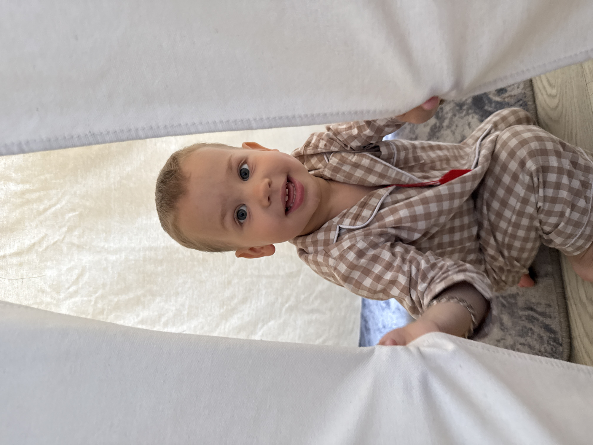 A toddler wearing brown and white checkered pajamas is sitting partially under a white sheet or blanket. The location is likely indoors, possibly in a child's play area, due to the presence of a rug and the playful context of the scene. The toddler is looking up with a smile, indicating engagement with someone or something outside the frame. Location: Rusanovo, Russia.