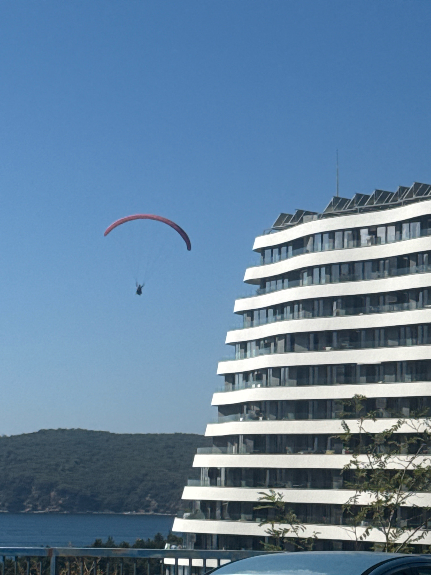 In Boreti, Montenegro, a paraglider is pictured against a clear blue sky, drifting over a body of water. To the right, a modern, multi-story building with a white facade and curved balconies dominates the scene. The building's design features staggered layers and numerous windows that reflect the sky. The water's edge is visible, bordered by a densely forested hillside in the background. In the foreground, a railing and the top of a vehicle are partially visible, suggesting a viewpoint from a slightly elevated position.