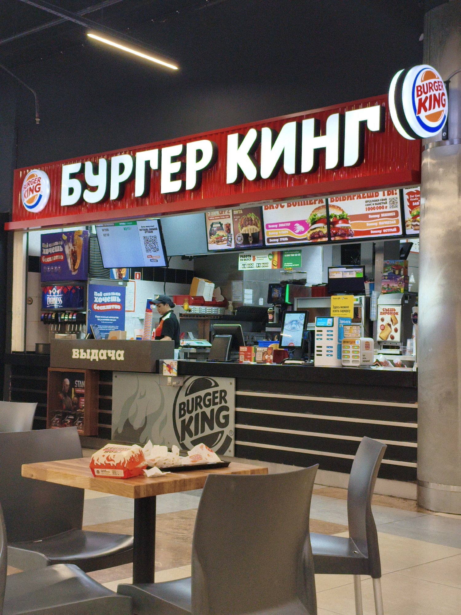 The image shows the interior of a Burger King restaurant. A large red sign with white lettering in Cyrillic, reading "БУРГЕР КИНГ" (Burger King), is prominently displayed above the counter. To the left of the sign, the familiar Burger King logo is visible. A food counter is set up with a digital menu board displaying prices and images of food items. A Burger King employee in uniform is seen behind the counter, interacting with a customer. Various menu displays and promotional materials are also visible. In the foreground, a table with chairs is set up, with a Burger King meal package and napkins on the table. The overall environment appears to be a typical fast-food restaurant setting within a commercial space.