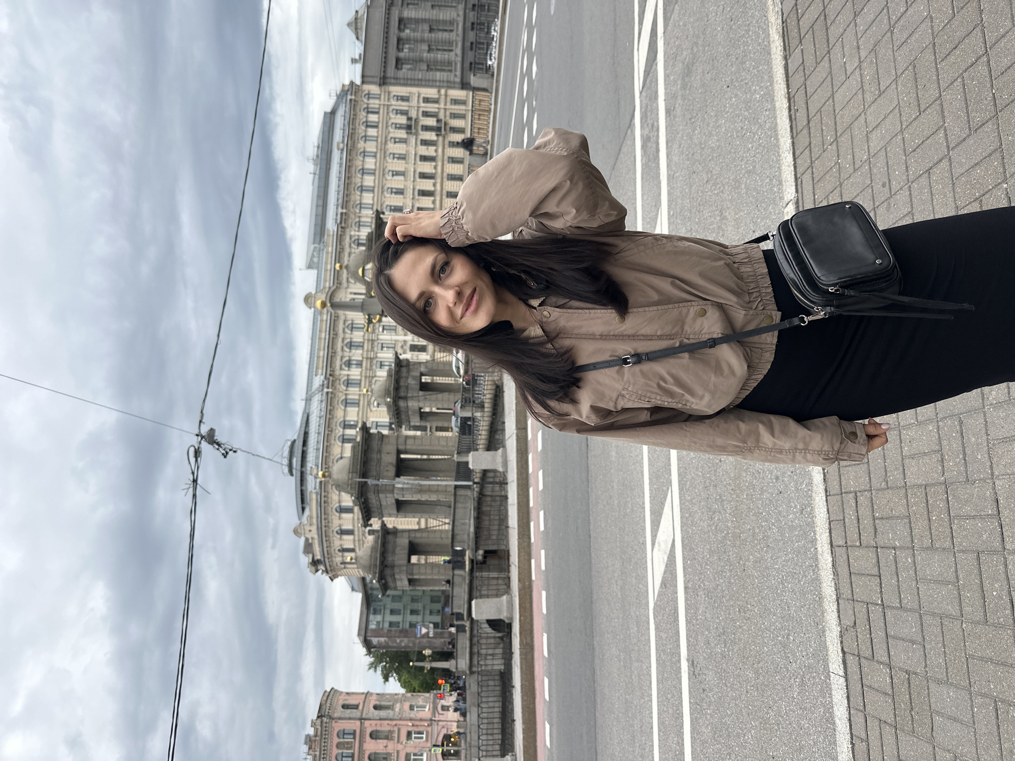 The image depicts a young woman with dark hair, wearing a brown cropped jacket and a black skirt, standing on a sidewalk. She is holding her hand to her hair and looking at the camera with a slight smile. A black cross-body bag hangs from her shoulder. Behind her, a street with white lane markings is visible, leading to a bridge with stone pillars and railings. Several buildings with classical architecture and a cloudy sky are in the background. The scene appears to be set in Saint Petersburg, Russia, given the architectural style of the buildings and the presence of a canal or river.