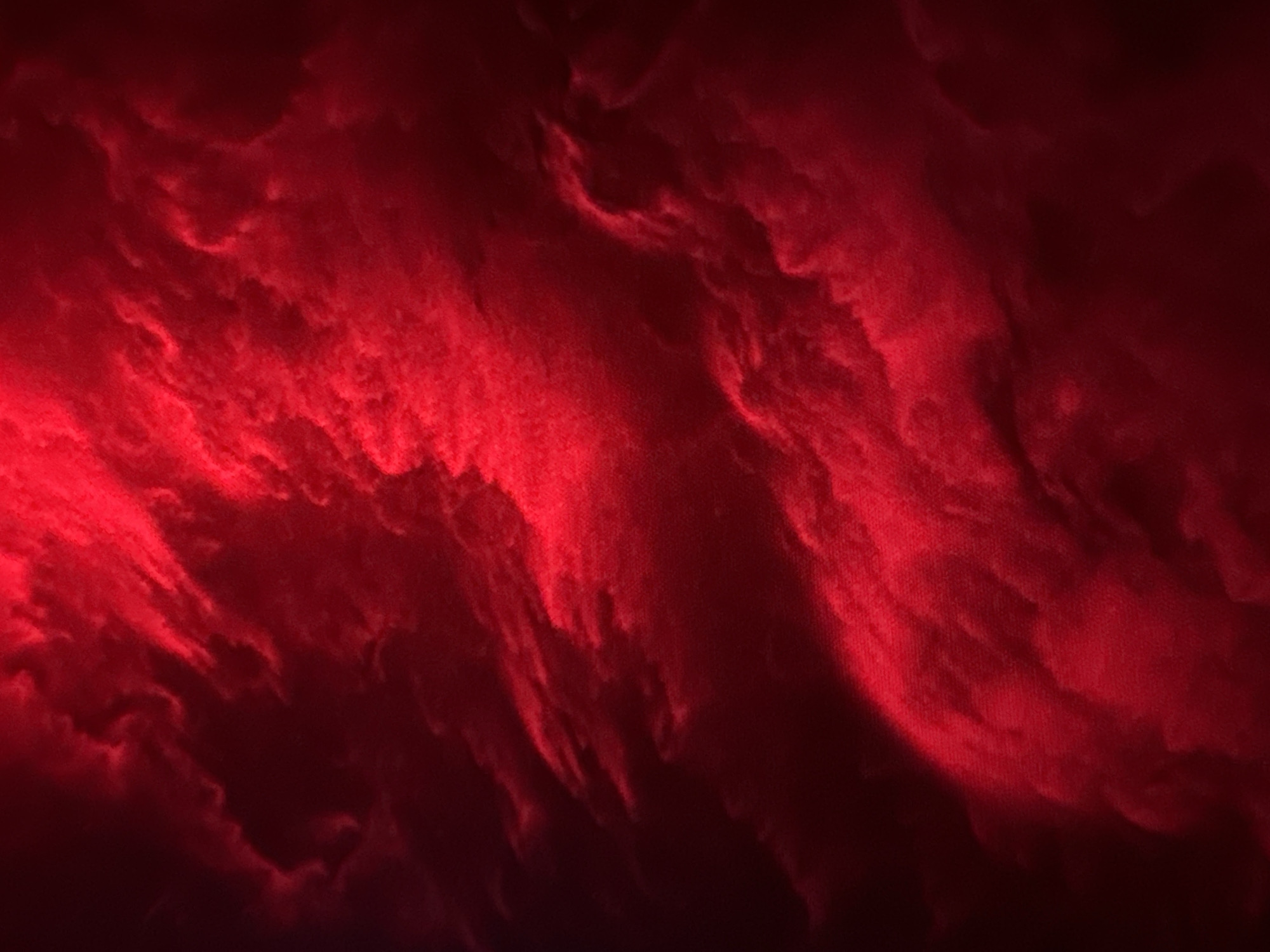 The image is a close-up of wispy, textured formations illuminated by a vibrant red light. The textures appear soft and organic, resembling smoke, clouds, or possibly a textured fabric. The lighting creates a dramatic contrast, with bright red areas and deep, dark shadows. The overall impression is abstract and atmospheric, with no discernible objects, people, or specific location details visible. The location is unknown.