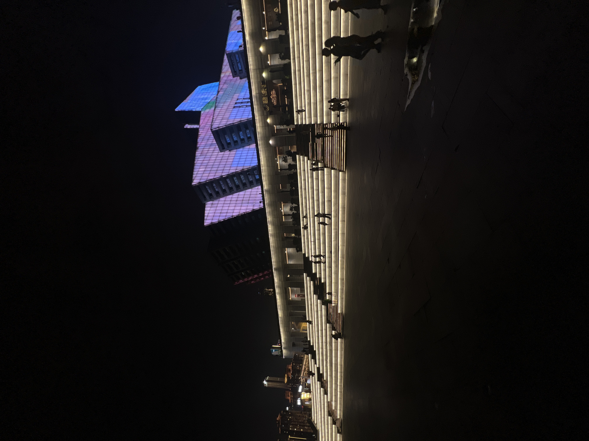 An exterior nighttime view of a wide, multi-tiered set of illuminated stairs leading up to a modern building. The building's facade is covered in a grid of lights that display vibrant pink, purple, and blue hues, giving it a dynamic, abstract appearance. Several people are visible on the stairs, some walking, others sitting or standing. In the foreground, silhouetted figures walk across a dark plaza, one of whom is near a puddle reflecting the building's lights. The city of 江北城街道, China, is visible in the background with illuminated skyscrapers.