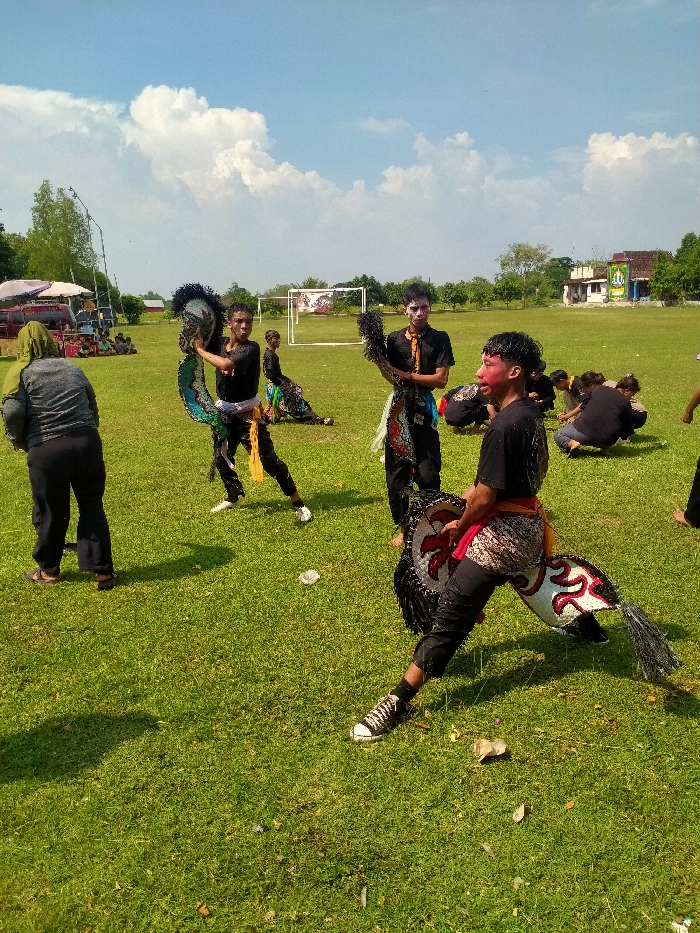 The image depicts an outdoor cultural performance or practice session on a sunny day in an open grassy field in Jetak, Indonesia. Approximately ten individuals are visible.
In the immediate foreground, a male performer is positioned on the right, in a deep squatting stance. He wears black sneakers, black pants, a black short-sleeved shirt, a red sash, and a patterned sarong. His face is painted with red and black accents. He holds a white and black Kuda Lumping (hobby horse) prop with red stylized patterns.
Mid-ground to the left, a male performer stands, holding a blue and black hobby horse prop. He wears white sneakers, black pants, a black short-sleeved shirt, and a yellow sash. His face appears unpainted, and he looks towards the viewer. Directly to his right, another male performer stands, holding a black feathered prop. He wears black pants, a black short-sleeved shirt, and a patterned sash. His face is painted predominantly white around the eyes. Further back on the left, a fourth performer is partially visible in a squatting position, similarly attired with a hobby horse prop.
In the far background on the right, several individuals are gathered, some squatting, others standing, likely additional performers or observers. On the far left foreground, an individual viewed from behind wears a gray long-sleeved top, dark pants, and a green head covering.
The environment consists of a vibrant green grass field, with minimal scattered white debris. A white metal soccer goal frame is visible in the distant center background. Trees and foliage line the field's edges, and in the far background to the right, a light-colored building with a blue and white mural or banner stands. To the far left background, a white tent structure with people gathered underneath is present. The sky is bright blue with scattered white cumulus clouds, indicating strong natural lighting.