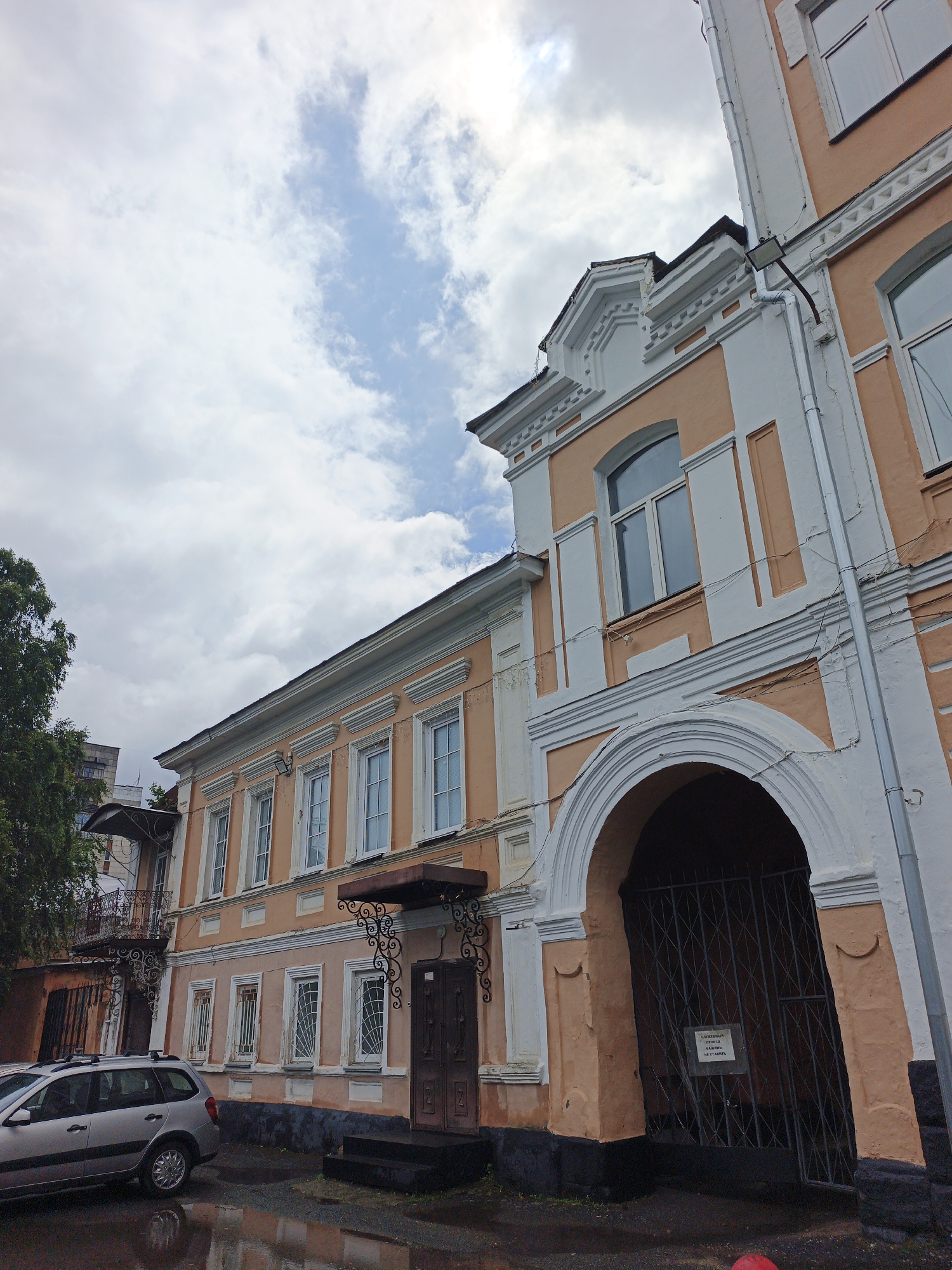 The image depicts a two-story building with peach-colored walls and white decorative trim. The building features a large arched entryway with a black metal gate. Above the gate, a sign with Russian text is visible. The building has multiple windows, some with grates. To the left of the building, a silver wagon-like car is parked on a wet asphalt surface, reflecting the surrounding environment. A large tree with green foliage is also present to the left, partially obscuring a taller, lighter-colored building in the background. The sky is cloudy with patches of blue. The scene appears to be an urban setting, possibly a residential or commercial area. The architectural style suggests an older building. The presence of the car and the wet ground might indicate recent rainfall. Based on the available information, this scene is located in the city of Zlatoust, Russia.