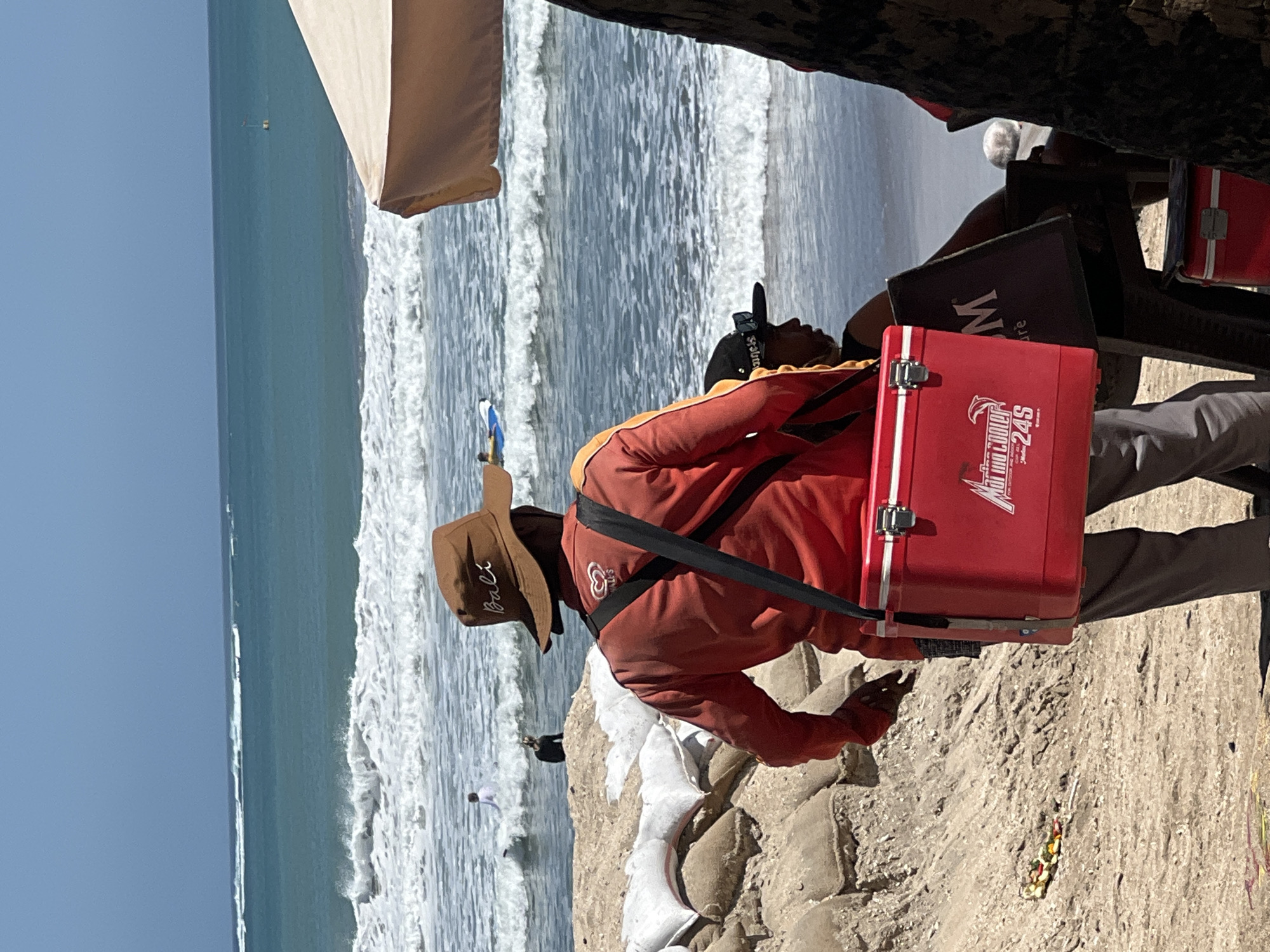 This image captures a scene on the sandy beach of Kuta, Indonesia, near the ocean. In the foreground, a person wearing a brown hat with "Bali" written on it and an orange long-sleeved shirt is seen from behind. They are carrying a red cooler bag with a black strap over their shoulder. The cooler bag has "Marino Cooler 24S" and a logo on it. In the middle ground, the ocean displays white waves breaking near the shore. Further in the background, two individuals are visible in the water; one is standing and another is sitting. Sandbags are piled up to the left of the person with the cooler. To the right, part of a light-colored umbrella and a tree trunk are visible. The overall environment is sunny with a clear blue sky.
