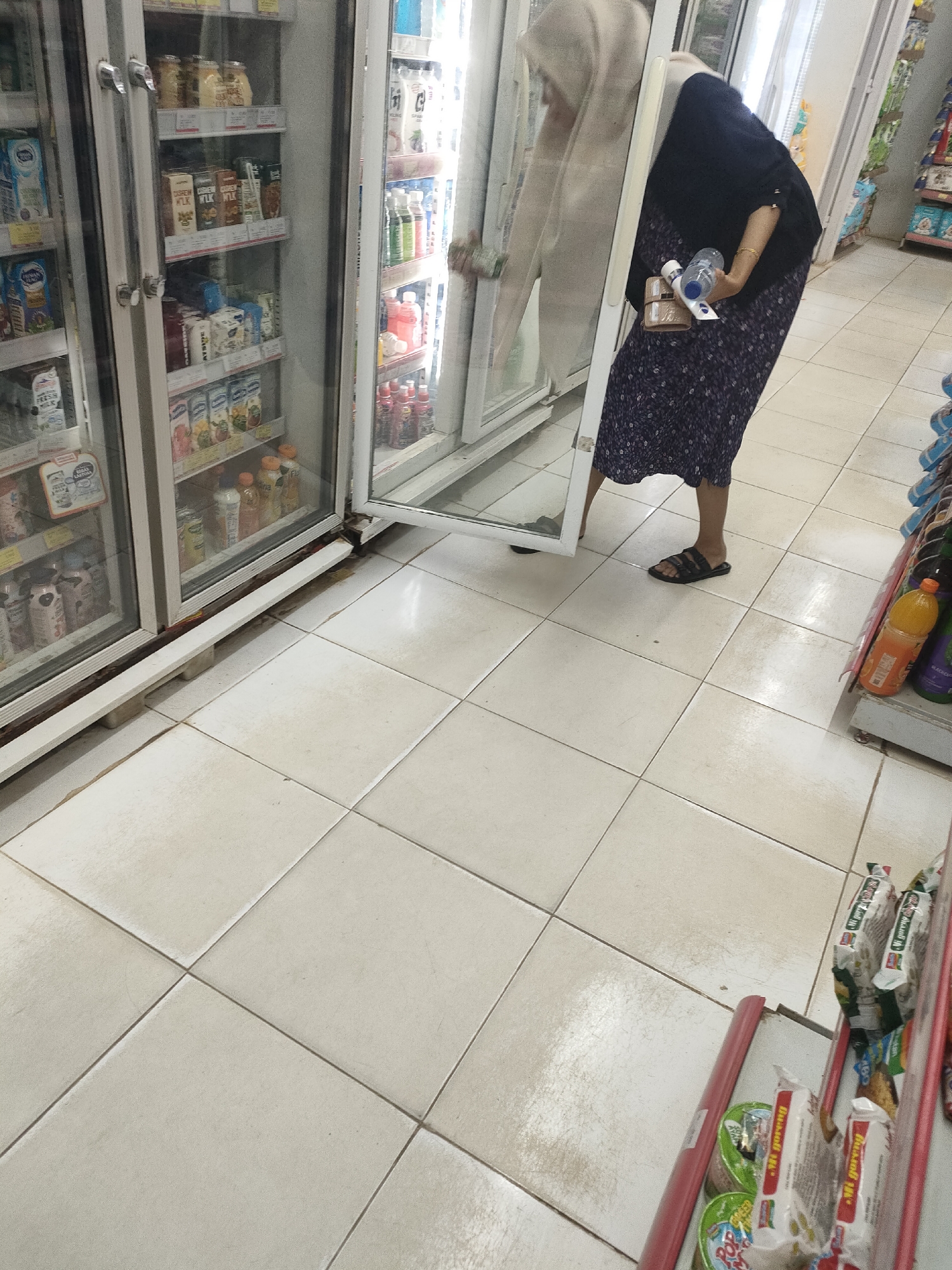 In Desa Banyumas, Indonesia, a woman wearing a dark blue hijab and a patterned dress is seen reaching into an open glass-door refrigerator. She holds a clear plastic water bottle in her right hand and a brown wallet in her left. The refrigerator's interior displays various packaged drinks and food items. The floor is tiled with off-white squares. To the right, shelves are stocked with bottled beverages, including one with an orange liquid and a yellow cap.