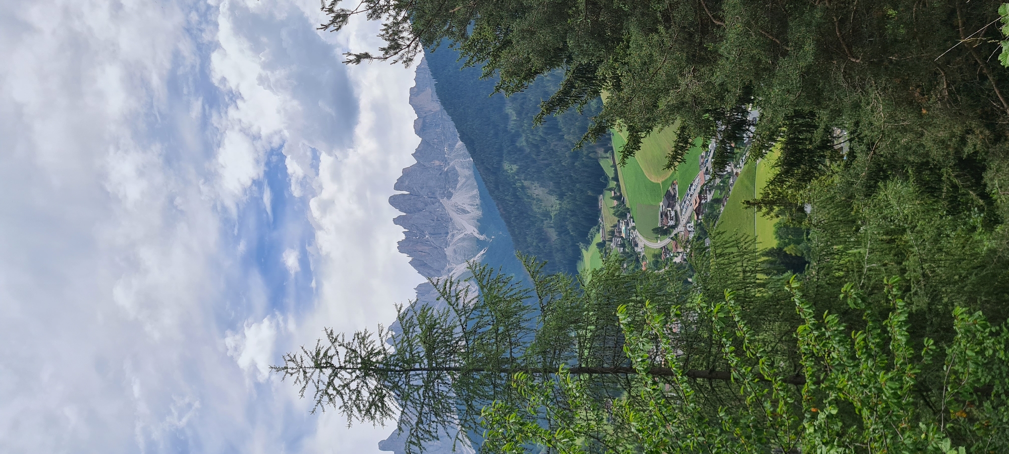 The image depicts a panoramic view of St. Magdalena, Santa Maddalena, Italy, nestled within a valley beneath jagged mountain peaks. In the foreground, dense evergreen trees frame the scene, with one prominent tree on the left dominating the lower portion of the frame. A winding road is visible leading into the village, which is characterized by traditional alpine architecture with red-tiled roofs. The village is surrounded by lush green fields, some of which are cultivated. In the background, a dramatic range of grey, rocky mountains rises sharply, their peaks partially obscured by dramatic, white, and grey clouds against a pale blue sky. The overall impression is one of a serene and picturesque alpine landscape.
