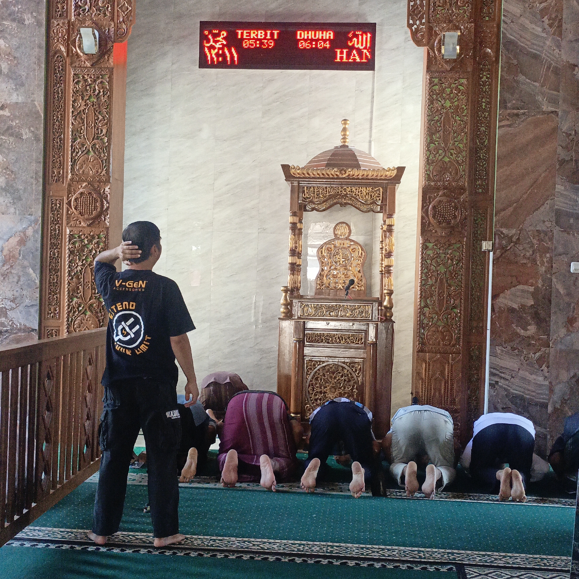 The image shows a group of men praying inside a mosque. The mosque appears to be decorated with ornate wooden carvings on the pillars and a wooden pulpit. A digital clock displaying prayer times is visible on the wall. One man, facing away from the camera, is wearing a black t-shirt with a logo and dark pants. The other men are in the foreground, bowed in prayer on a green carpet. The overall atmosphere suggests a solemn religious gathering. The mosque's design, particularly the carvings and the digital prayer time display, is typical of mosques in urban Indonesia. While the exact location is not specified in the image, such mosques are common in cities like Salatiga, Indonesia.