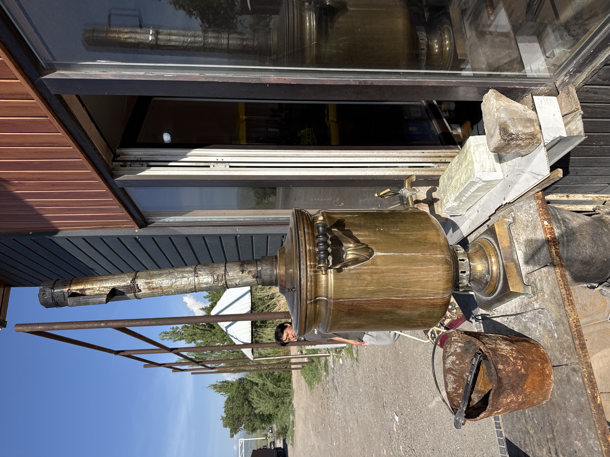 This outdoor scene, captured in Sasyk-Bulak, Kyrgyzstan, features a large, ornate brass samovar as its central subject. The samovar, a traditional tea urn, has a tall, galvanized metal stovepipe extending upwards from its top and a spigot on its side. It rests on a square base on what appears to be an outdoor workstation or platform.
To the right of the samovar, a rusty metal bucket with a handle sits on the ground, containing a dark tool resembling tongs. Behind the samovar, a young person with dark hair, wearing a grey t-shirt and grey pants, is partially visible, looking towards the camera.
The background shows an expanse of dirt ground or a road, with scattered green trees under a clear, bright blue sky with a few wispy clouds. A metal framework, possibly part of an awning or pergola, stretches across the middle ground. To the left, a building with reddish-brown horizontal wooden siding features large glass windows. These windows reflect parts of the outdoor environment and some indistinct shapes inside, but no clear text is visible. The scene is brightly lit by natural sunlight, suggesting a clear day, likely midday given the strong shadows. The overall setting is rustic and appears to be an operational outdoor area, possibly for serving hot beverages.