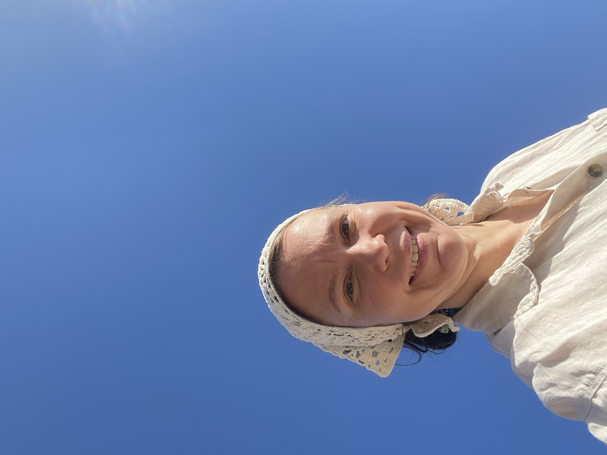 A low-angle, rotated view captures a woman against a cloudless, vibrant blue sky. She wears a light-colored linen shirt with buttons and a white, crocheted bonnet or head covering. She is smiling, and the sun casts light across her face. The location is not discernible in the image, and there is no basis to determine that it is Chaikovsky, Russia.