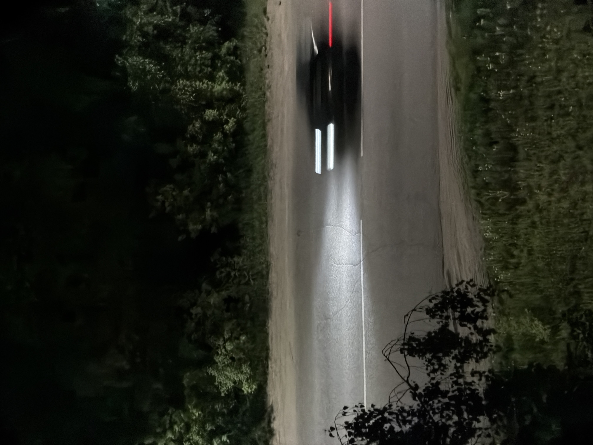 The image shows a dark road at night in what appears to be a rural area. A car is traveling on the road, its headlights creating bright streaks and its taillights creating a red streak. The road is surrounded by dark vegetation, including trees and bushes. The exact location in the city of Chaikovsky, Russia is not clear from the image.