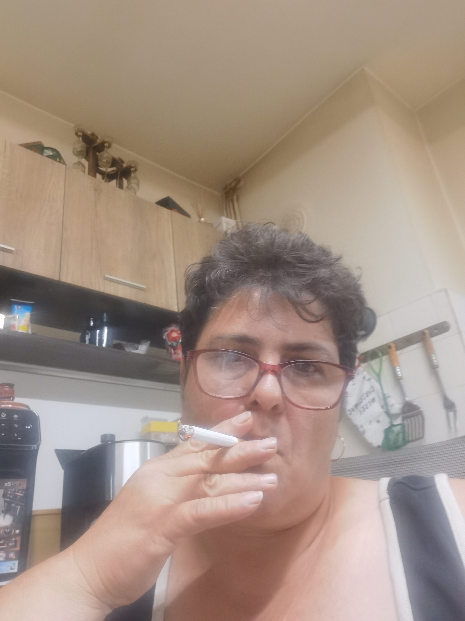 In a dimly lit interior, a person with short, curly, dark hair and glasses is captured in a close-up shot. They are holding a lit cigarette to their lips, with the tip of the cigarette and the ash clearly visible. The person is wearing a dark top with white trim. The background reveals kitchen cabinetry, shelves with various items, and cooking utensils hanging on a wall. The overall setting suggests a domestic environment, possibly a kitchen. The image does not provide any information regarding the city of Lomets, Bulgaria.