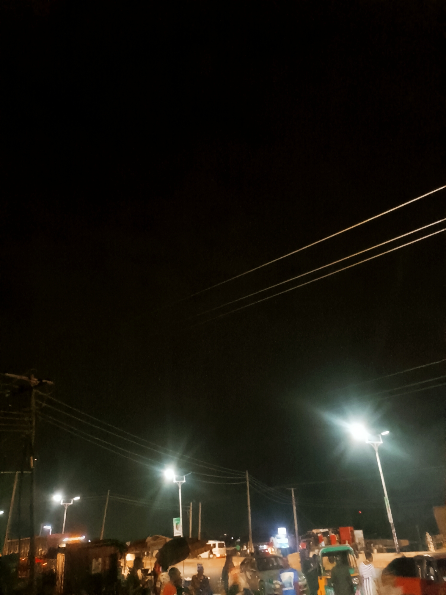 The image depicts a night scene in Bukuru, Nigeria. Streetlights illuminate a roadside area with various people, vehicles, and informal structures. People are seen standing and interacting near the road, possibly engaged in commercial activity or waiting for transport. Vehicles, including cars and three-wheeled auto-rickshaws (tuk-tuks), are visible on the road. There are rudimentary structures or stalls along the roadside, suggesting a market or trading area. Power lines are visible overhead. The overall scene suggests a bustling night-time environment.