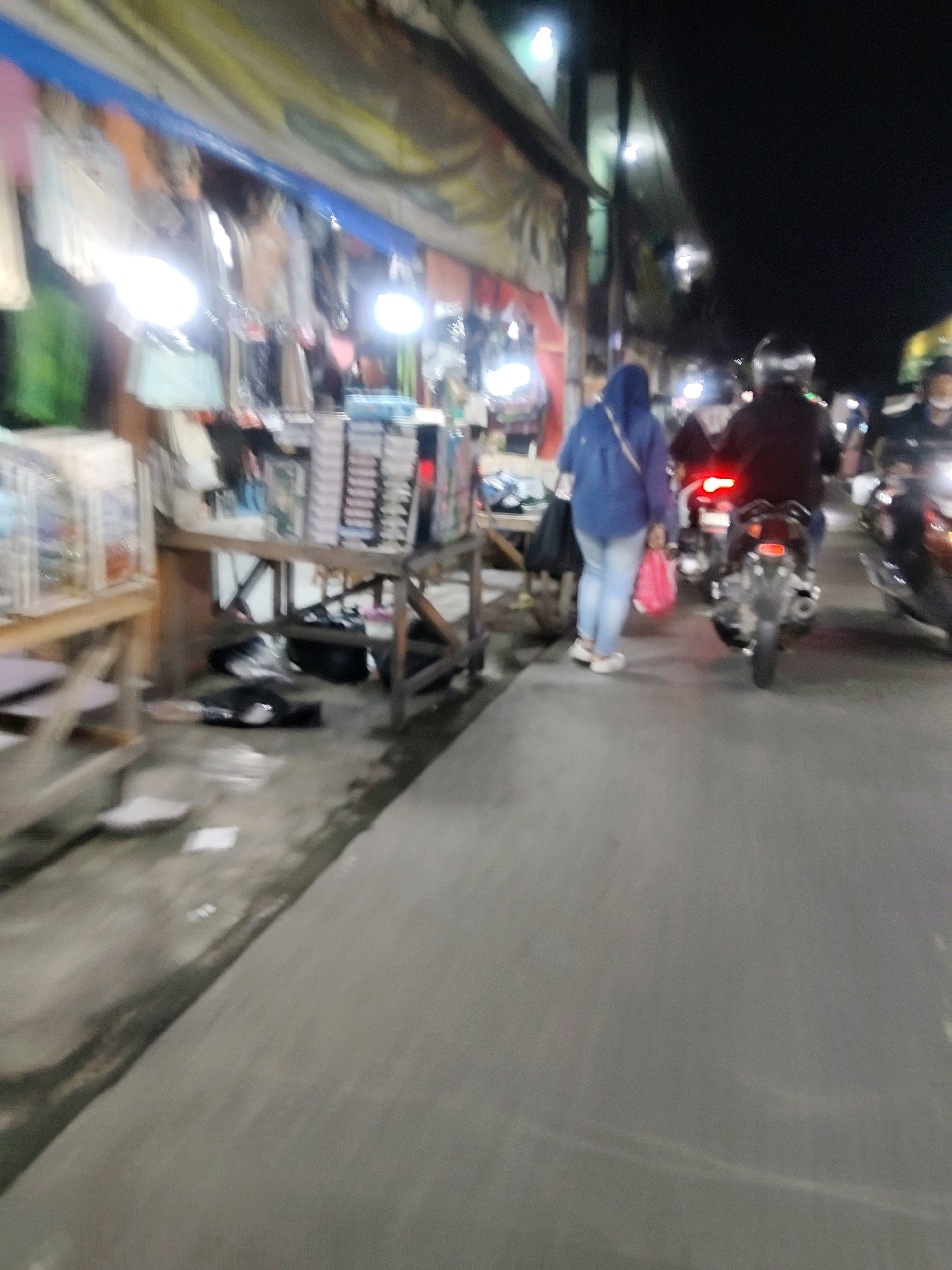 The image captures a nighttime scene on a street in Mekarmukti, Indonesia, likely a market area. In the foreground, a grey concrete road stretches into the distance. To the left, a market stall displays various items, possibly clothing, under bright artificial lights. Merchandise is hung from racks and shelves. On the right side of the road, several motorcycles are present, with riders on some of them. One motorcycle has its brake light illuminated. A person wearing a blue hijab and light-colored pants is walking away from the camera, carrying a pink bag. Another person is seated on a motorcycle behind the individual in the blue hijab. The overall lighting suggests it's evening or night, with streetlights illuminating the market stalls and the road. The ground appears somewhat cluttered with debris.