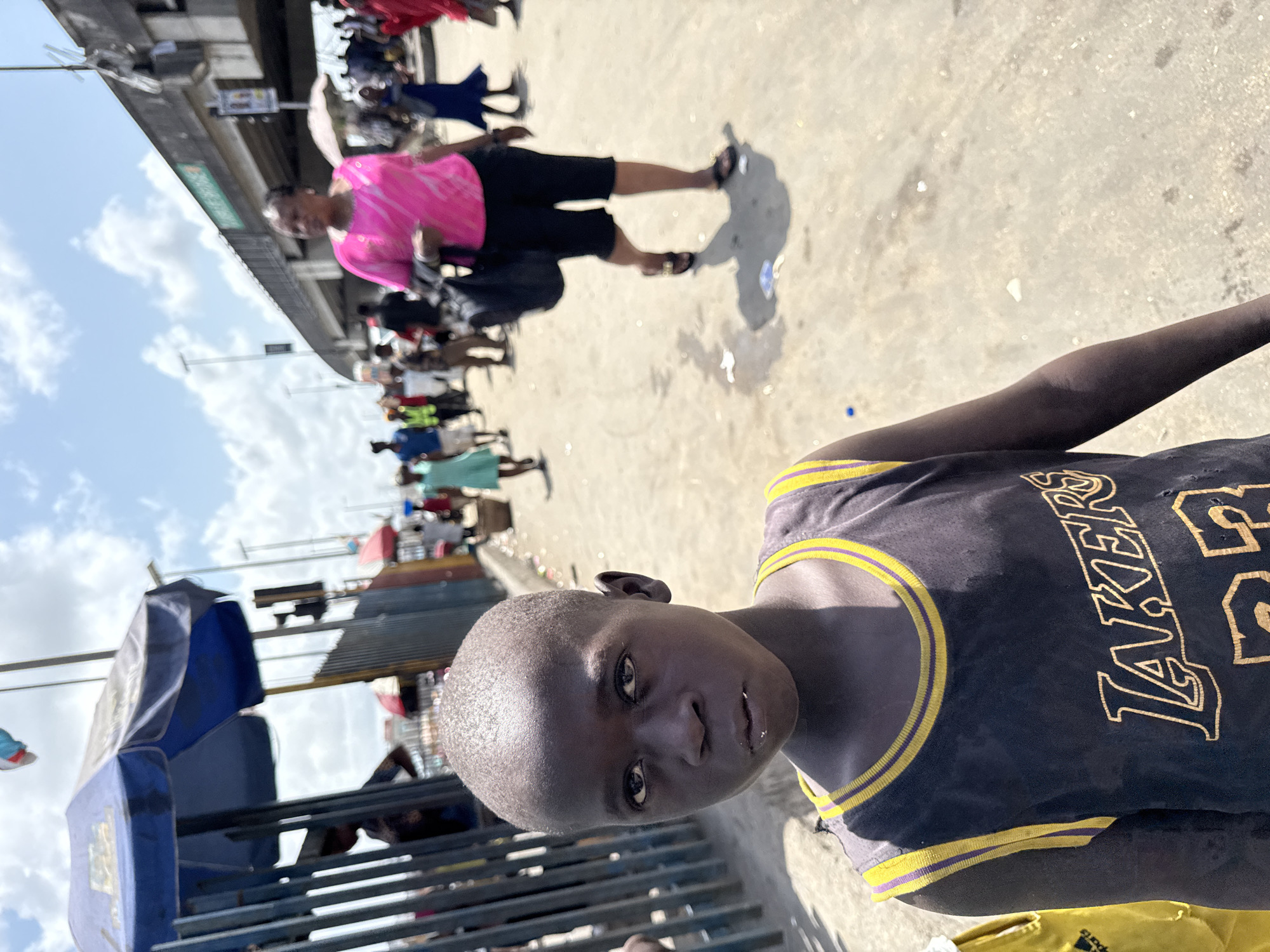 The photo appears to be taken in Lagos, Nigeria. In the foreground is a dark-skinned youth wearing a Lakers basketball jersey. Behind the youth, there is a crowded outdoor market scene with multiple people walking and standing under temporary shelters and metal structures. The background shows a mix of concrete structures, including what looks like an elevated walkway or bridge, and a partly cloudy sky. One person in a pink shirt and black shorts is visible walking away from the camera in the middle ground.