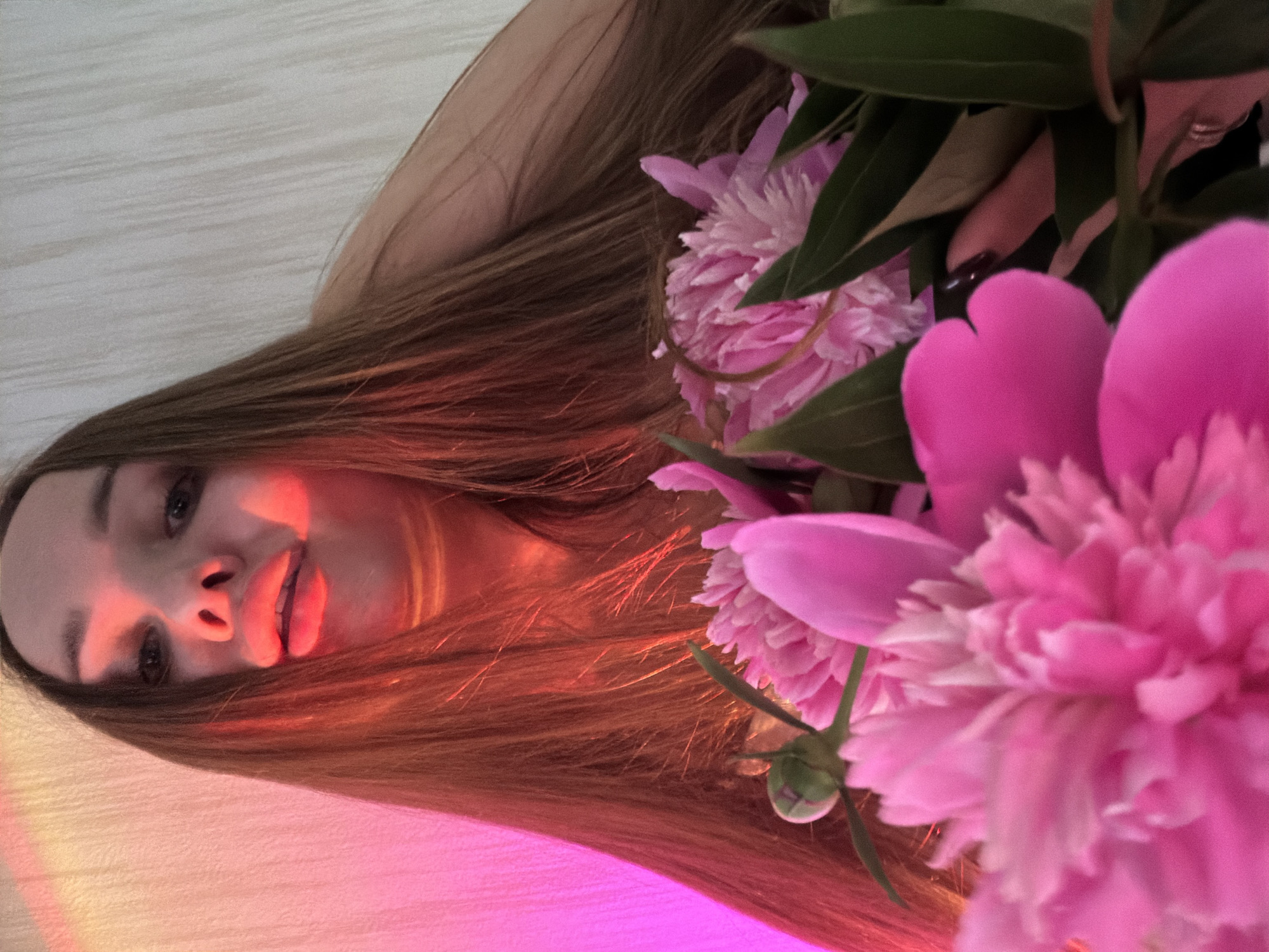 The image shows a young woman from a low angle, holding a bouquet of bright pink peonies in front of her face. The woman has long, brown hair and her mouth is open, with her lips slightly parted. She is looking directly at the camera. The background is a textured wall, illuminated by a pinkish-orange light, creating a dramatic and warm atmosphere. The peonies are the primary focus in the foreground, with their lush petals and green leaves taking up a significant portion of the frame. The image appears to have been taken indoors, likely in a residential setting. There is no information in the image that suggests a specific location in Chaikovsky, Russia.