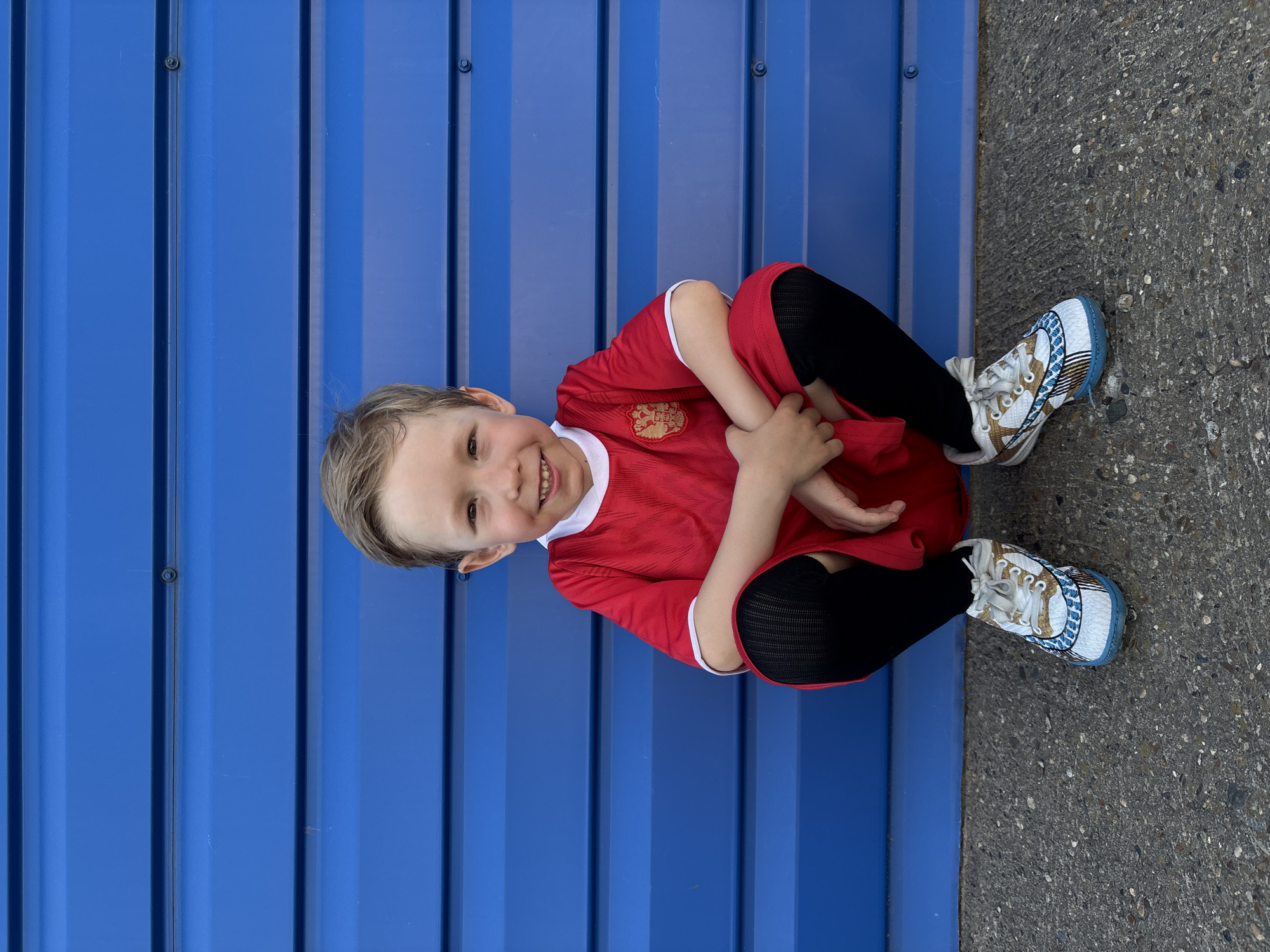 A young boy sits on the ground, leaning against a corrugated blue metal wall. He is wearing a red shirt with a white collar, black leggings, and patterned sneakers. The ground to his right is concrete. The location is in the city of Chaikovsky, Russia. The boy is smiling.