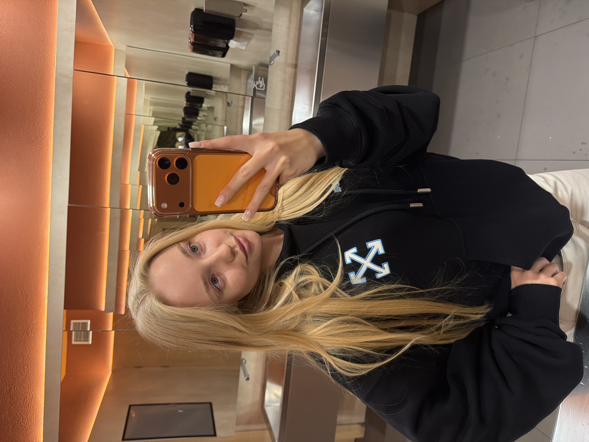 A young woman is taking a selfie in a public restroom. The woman has long blonde hair and is wearing a black hoodie with a white and blue design. She is holding a smartphone with a tan case. The restroom has an orange accent wall and multiple mirrors. There are sinks and paper towel dispensers visible in the background. The specific location within the city of Unknown, Unknown is not evident in the image.