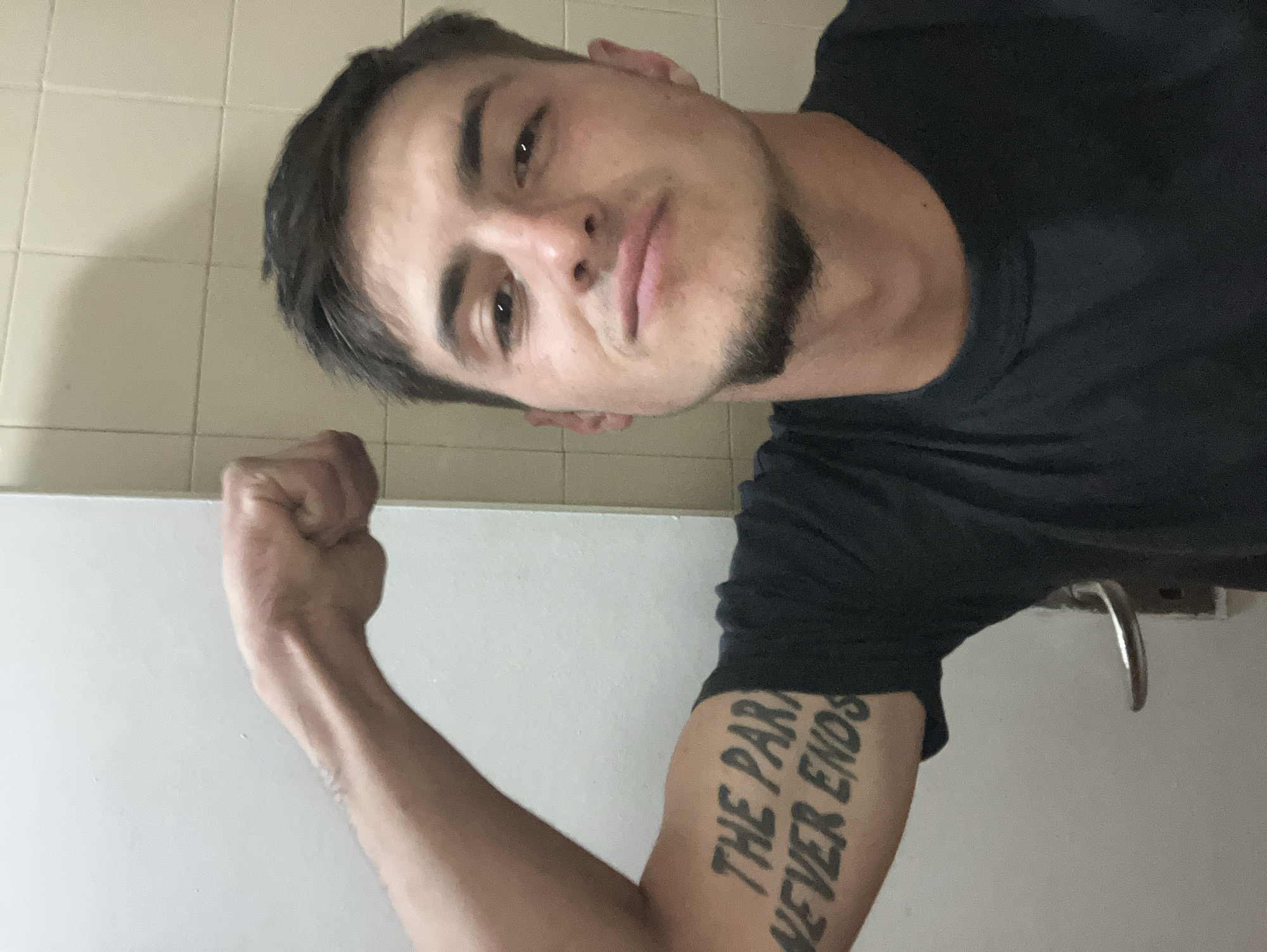 A man poses for a selfie, flexing his left arm. The tattoo on his arm reads "THE PARTY NEVER ENDS" in a stylized font. He is wearing a dark t-shirt. The background appears to be a tiled wall, possibly a bathroom. A metal handle is visible on the wall behind him. The location in the city of Plovdiv, Bulgaria, is not discernible from the visual information provided.