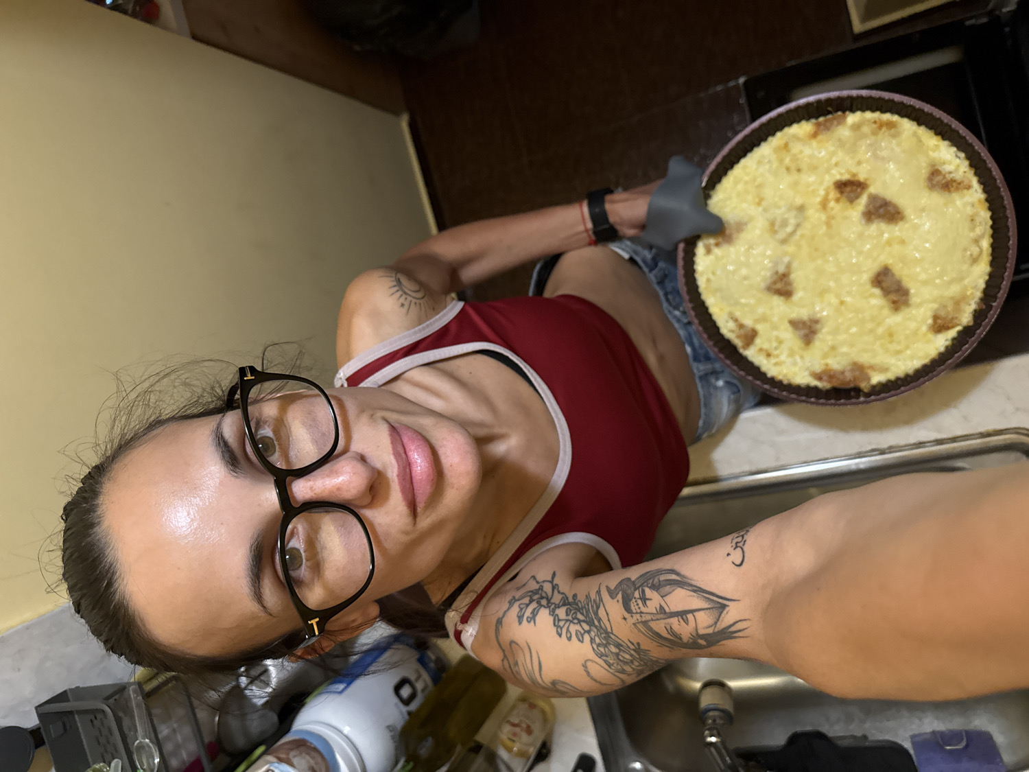 A young woman, likely in her 20s or 30s, is captured in a high-angle, somewhat tilted selfie or photograph within a kitchen setting in Pleven, Bulgaria. She is looking directly up at the camera with a neutral to slightly smiling expression. Her dark hair is pulled back, and she wears black-framed glasses with a visible "T" logo on the side. She is dressed in a red tank top with white trim and light-colored denim shorts.
Her left hand, protected by a grey oven mitt, holds up a round baking dish or pan. The dish contains a light yellow, textured food item, possibly a casserole or baked dessert, with several browned, crusty pieces on its surface, indicating it has recently been baked.
Visible tattoos include a sun and moon design on her left shoulder, a larger, detailed graphic (appearing to be an eye or mask) on her right arm, and a smaller, stylized symbol (resembling a "3") on her right inner elbow. Her skin appears glossy or dewy.
The background shows a home kitchen environment, with a stainless steel sink and faucet visible in the foreground to her right. A dark-colored countertop stretches behind her, cluttered with various bottles, containers, and possibly kitchen appliances or parts. A light, warm yellow wall is seen to her left, and a dark oven or cabinet is in the far background. The overall scene suggests a moment after preparing or baking food.