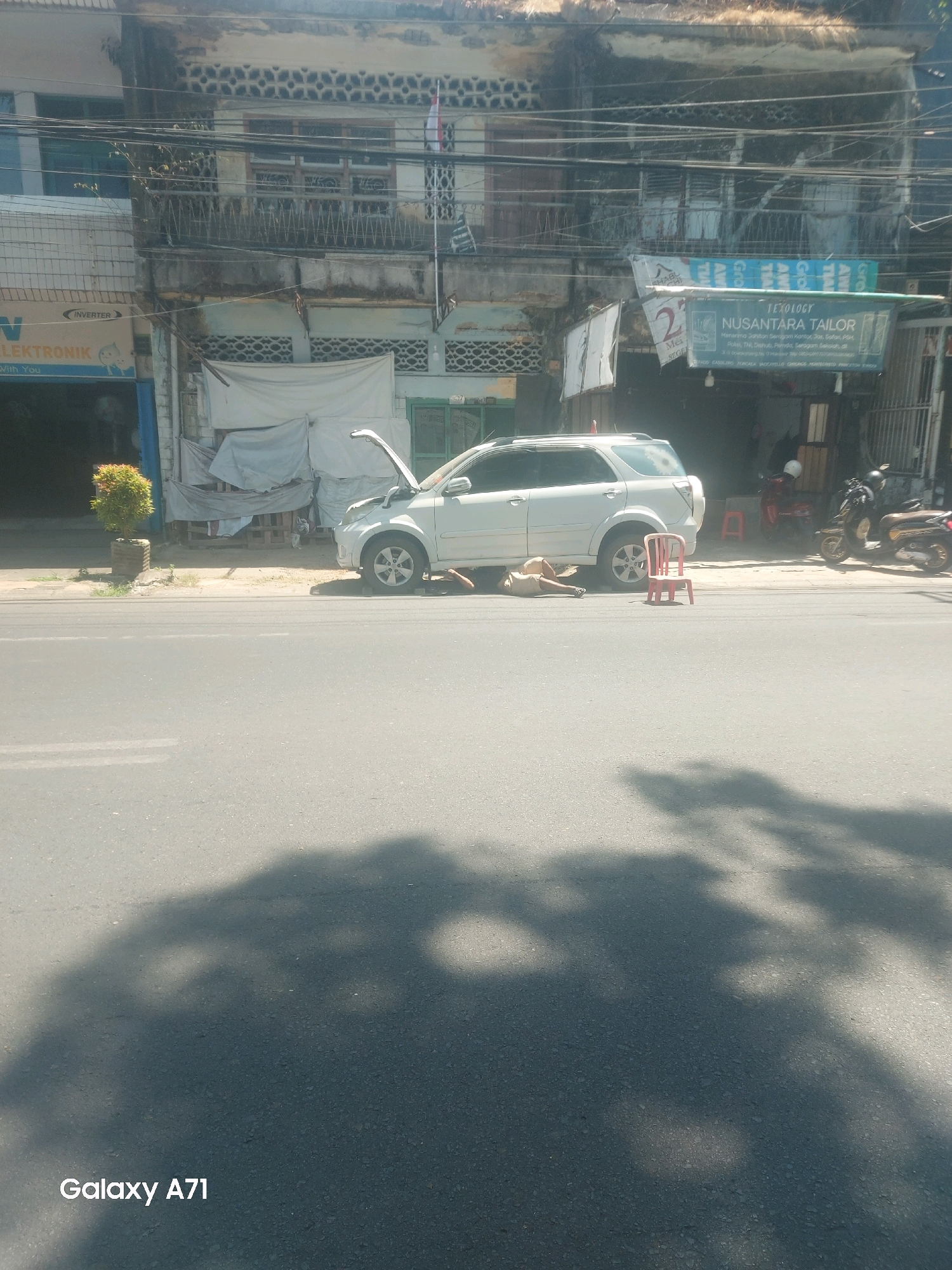 The image depicts a street scene in Makassar, Indonesia. A white SUV with its hood open is parked on the side of the road. In front of the SUV, a person is lying on the ground, appearing to be asleep or resting. A small red plastic chair is placed next to the vehicle. In the background, a two-story building with several shops and businesses is visible, including one with a sign that reads "NUSANTARA TAILOR." Several motorcycles are parked in front of the building. The scene is bathed in sunlight, casting shadows on the asphalt road.