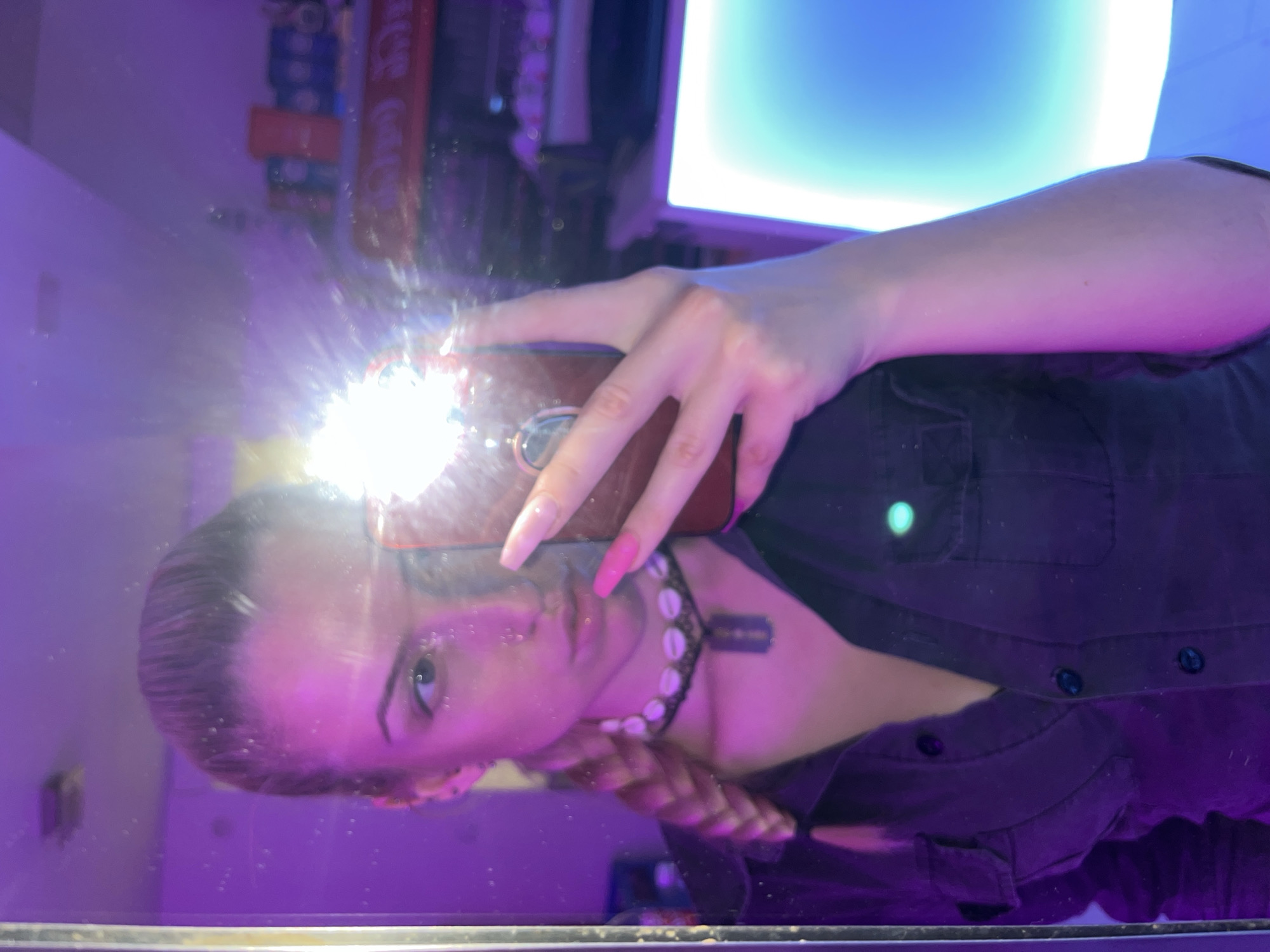 A person is taking a selfie in a mirror. The person is holding a phone with the flash on, obscuring part of their face. The person is wearing a dark button-down shirt and a necklace. The background is blurry but shows items on a shelf, including a Coca-Cola can. The location is unknown and there is no definitive evidence in the image to suggest it is in Rakovski, Bulgaria.