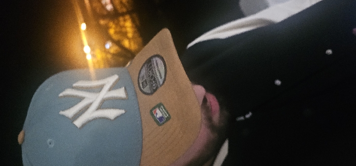 A person wears a light blue New York Yankees baseball cap with a tan brim. The cap features a white "NY" logo on the front. The brim of the hat also features stickers. The person is also wearing a black varsity jacket with white details. The image is grainy and taken in dim lighting.