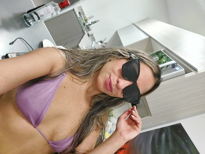 An adult female subject is centered in a medium close-up, appearing to take a selfie indoors. She wears a light purple, glittery bikini top and large black-framed sunglasses, which she is adjusting with her right hand. Her long, light-brown hair with blonde highlights frames her face. A silver philtrum piercing is visible above her upper lip.
The background indicates an indoor setting, likely a modern kitchen or dining area in a private residence. To the subject's left, a kitchen area features a stainless steel sink, a chrome faucet, and a light-colored granite countertop. Gray base cabinets and light-colored upper cabinets are visible. A white appliance, possibly a microwave, houses or displays a green potted plant. To the subject's right, a portion of a wall displays an art piece depicting a dark, starry sky above a landscape with pine trees and an orange glow near the horizon.
In the foreground, on a dark, flat surface (possibly a kitchen island or table), a clear glass bottle with a red cap is present. The bottle's label contains text, including "Buenos Aires" and "Argentina," and a barcode. A partially visible white plate and a black cable with an electronic device (possibly a remote control) are also on the surface. The specific location within Buenos Aires, Argentina, beyond an indoor residential setting, cannot be determined from the provided image.