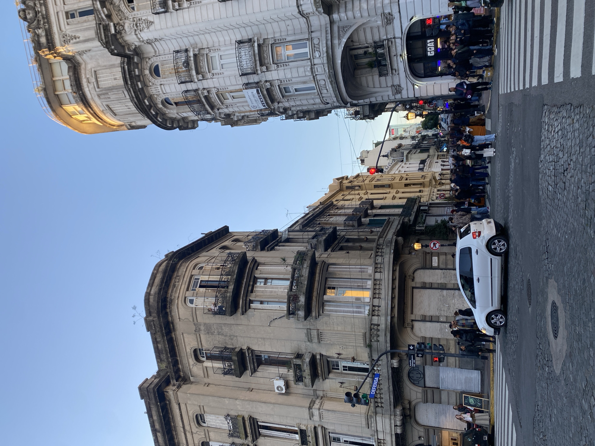 The image shows a street scene in Buenos Aires, Argentina. The view is angled upwards, showcasing the architecture and street-level activity. Two ornate buildings with European-style facades stand on opposite sides of the street. One building is white with elaborate decorations, while the other is a weathered grey, with multiple balconies and windows. A line of people waits to cross the street at a zebra crossing. A white car is parked at the side of the road. The sky is a clear, light blue. Street lights, traffic signals, and signage are visible. A GOAT logo is visible above a store entrance.